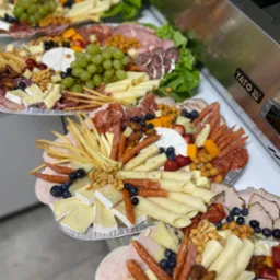 Kilka aluminiowych tac z różnorodnymi przekąskami: sery, wędliny, owoce (winogrona, borówki, truskawki) i orzechy ułożone kompozycyjnie na przyjęcie.