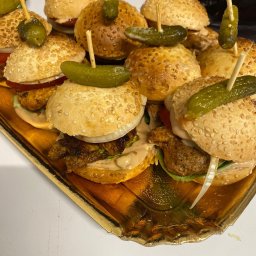 Adam Oberda - Food Group - Mini burgery z sezamową bułką, mięsem, warzywami i sosem, spięte wykałaczką z ogórkiem konserwowym, ułożone na złotej tacy.