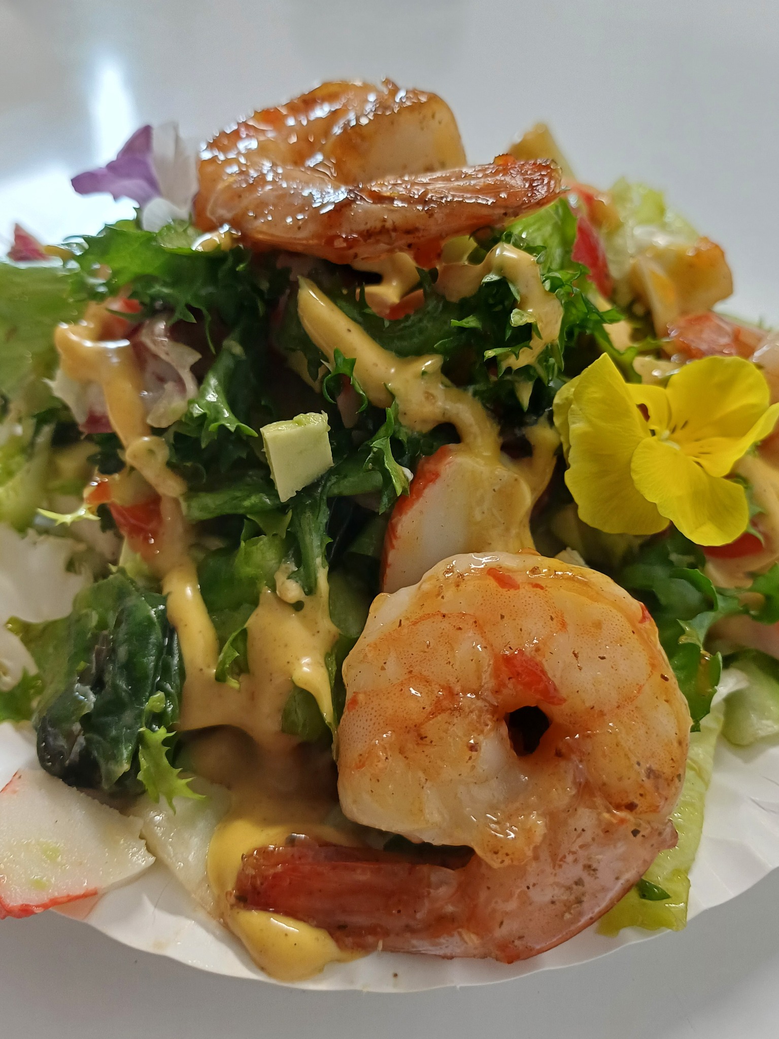 Apetyczna sałatka z krewetkami, zielonymi liśćmi sałaty, surimi, kawałkami awokado, polana kremowym sosem i udekorowana jadalnymi kwiatami, podana na białym talerzu.