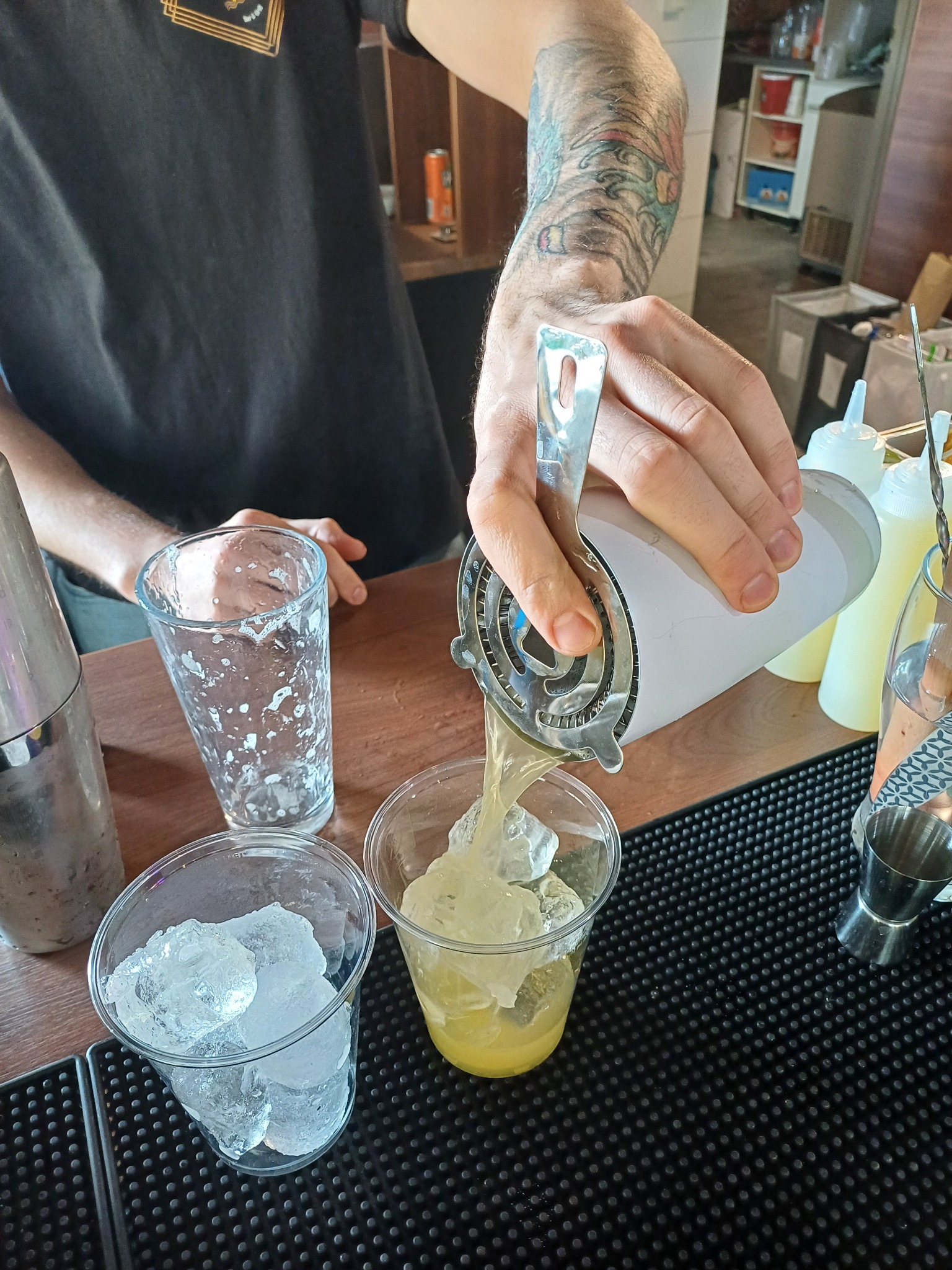 Barman z tatuażem na ramieniu przelewa przez sitko koktajl z shakera do plastikowego kubka z lodem na czarnej macie barmańskiej, obok miarka barmańska i puste naczynia.