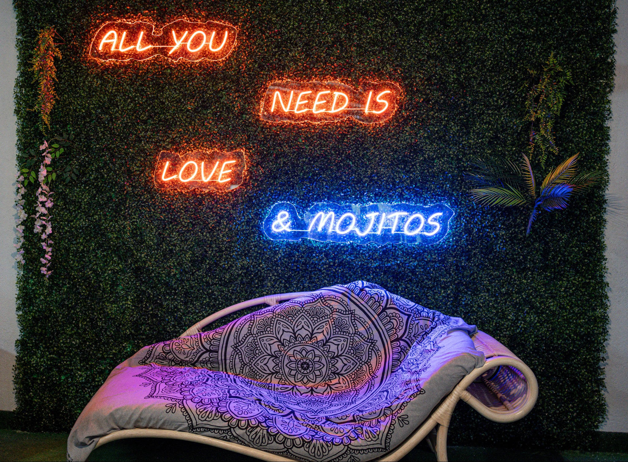 Rattanowy fotel z poduszkami w stylu boho, podświetlony neonowymi napisami 'All you need is love & mojitos' na tle ściany z zielonych roślin.