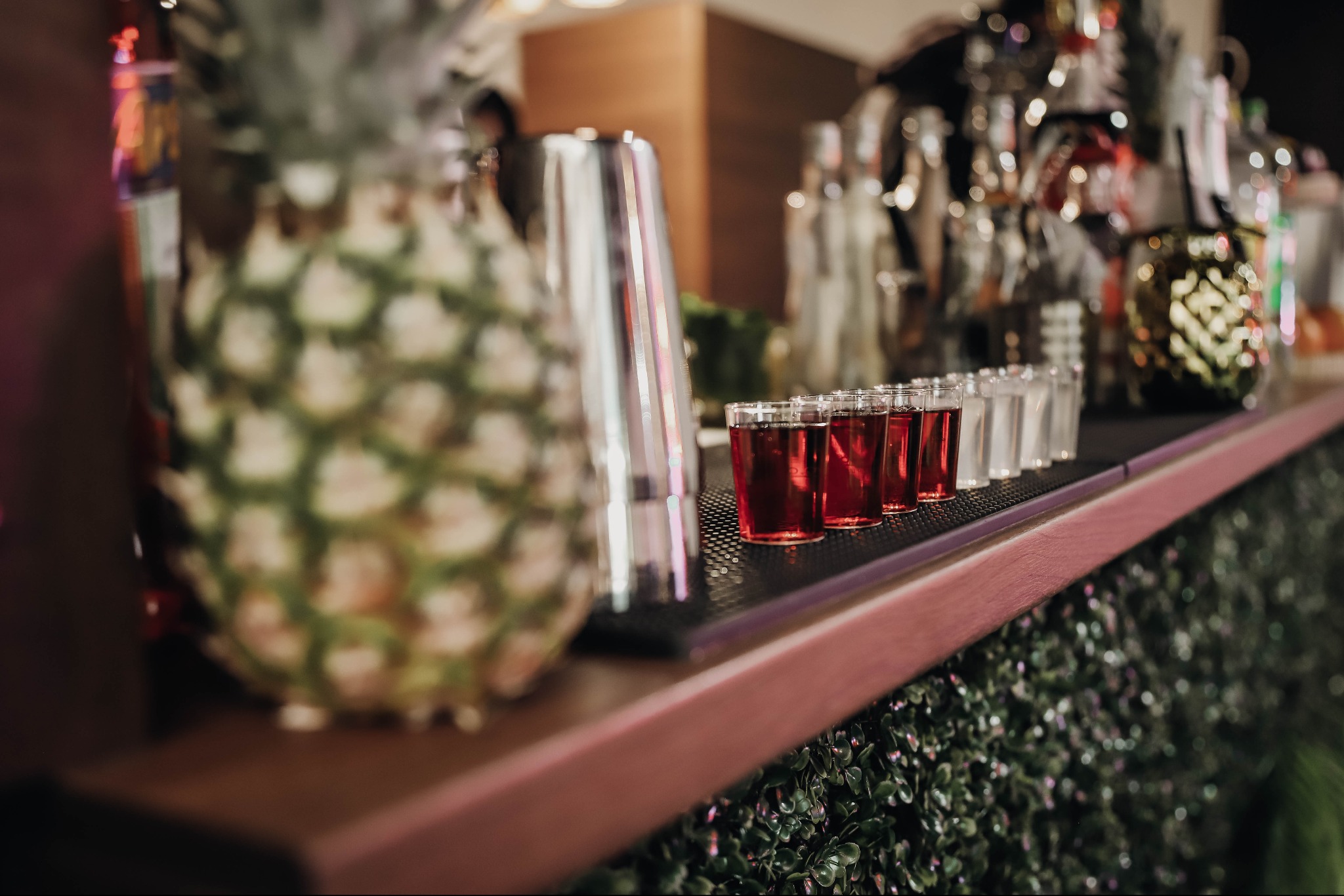 Bar z ananasem w tle, metalowym shakerem i rzędem kieliszków z czerwonym napojem, udekorowany zielonymi roślinami.