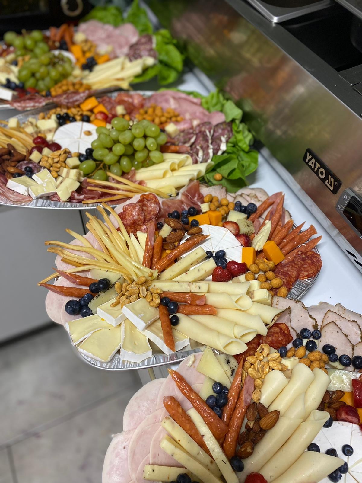 Kilka aluminiowych tac z różnorodnymi przekąskami: sery, wędliny, owoce (winogrona, borówki, truskawki) i orzechy ułożone kompozycyjnie na przyjęcie.