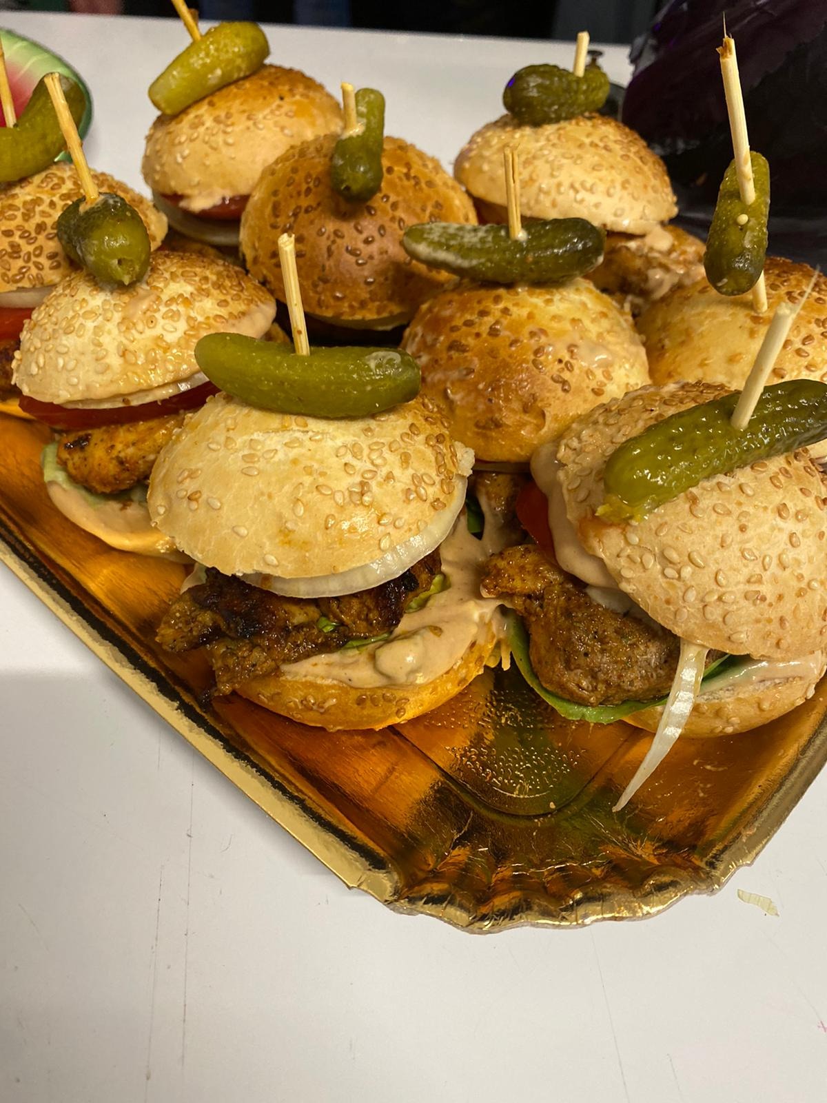 Mini burgery z sezamową bułką, mięsem, warzywami i sosem, spięte wykałaczką z ogórkiem konserwowym, ułożone na złotej tacy.