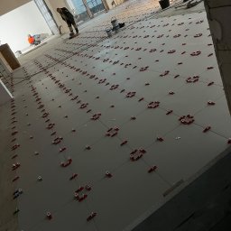 AirForce Wentylacja Krzysztof Chaciński - Układanie płytek podłogowych z użyciem klipsów poziomujących w Lublinie. Widoczni pracownicy i narzędzia glazurnicze. Duża powierzchnia, jasne płytki.