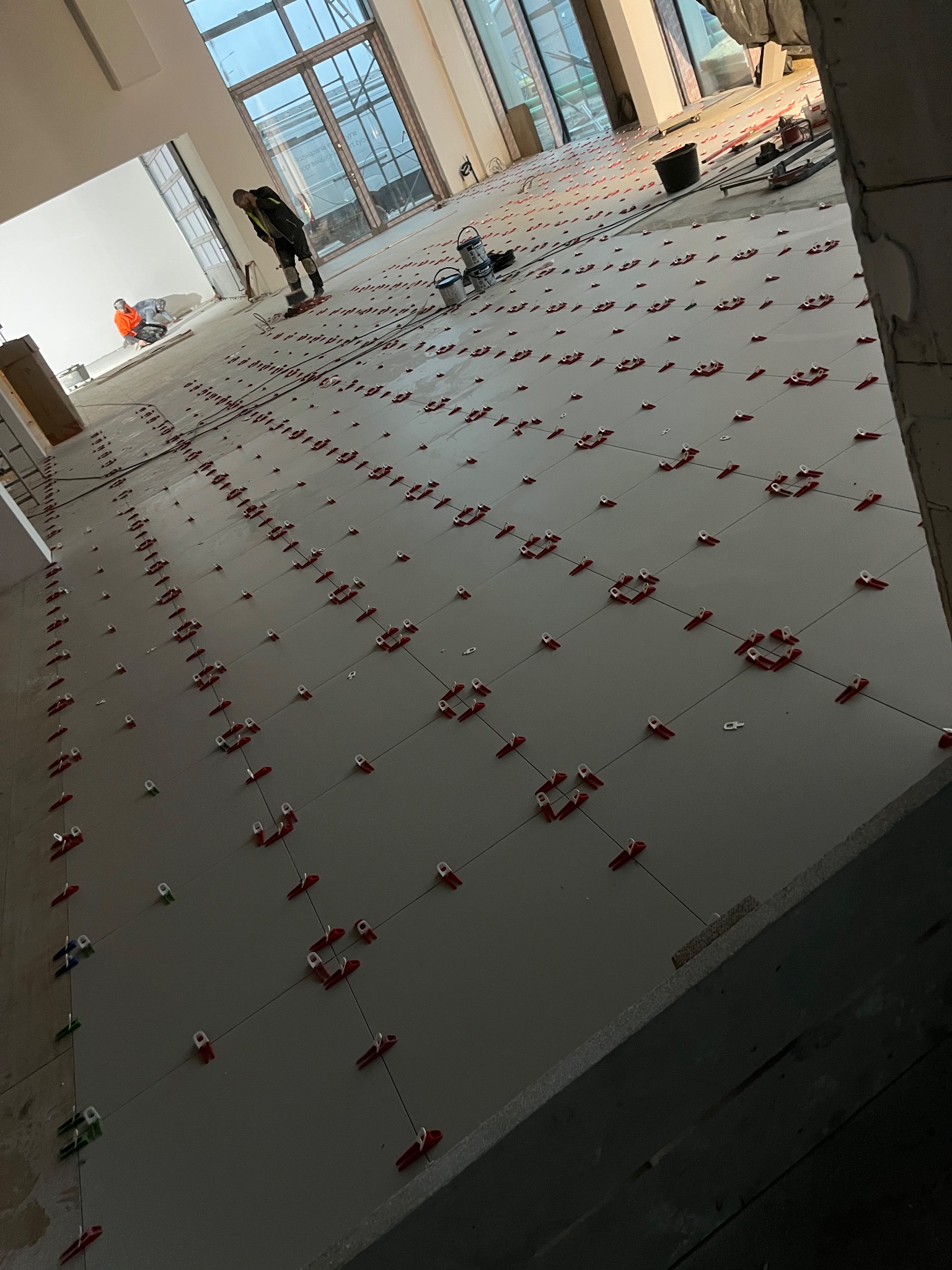 Układanie płytek podłogowych z użyciem klipsów poziomujących w Lublinie. Widoczni pracownicy i narzędzia glazurnicze. Duża powierzchnia, jasne płytki.