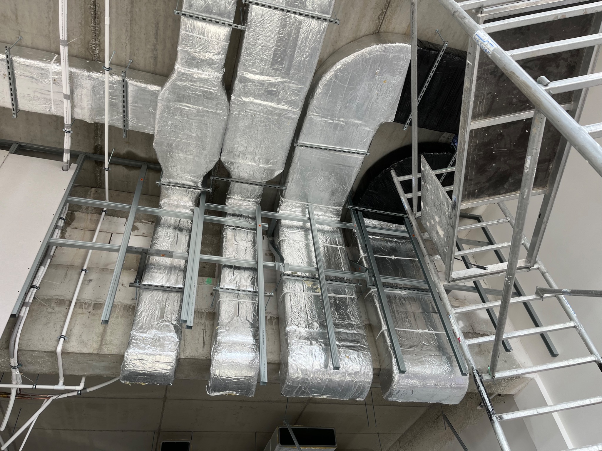 Srebrne, zaizolowane kanały wentylacyjne pod sufitem, zamontowane na metalowej konstrukcji. Widoczne rury i drabina. Ujęcie z dołu, szeroki kadr.