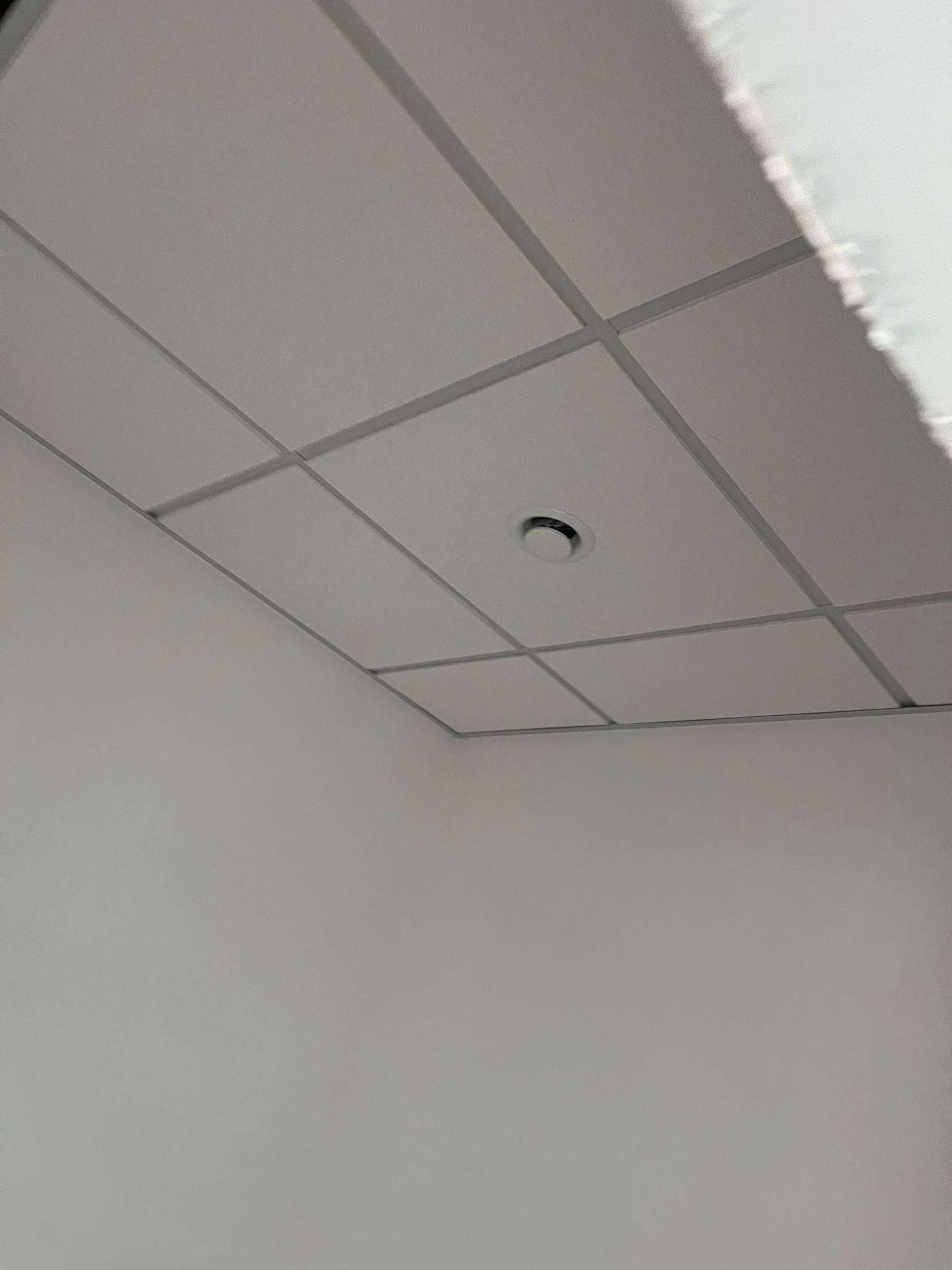 Biały sufit kasetonowy z okrągłym nawiewnikiem wentylacyjnym w rogu pomieszczenia. Minimalistyczny design, neutralne oświetlenie.