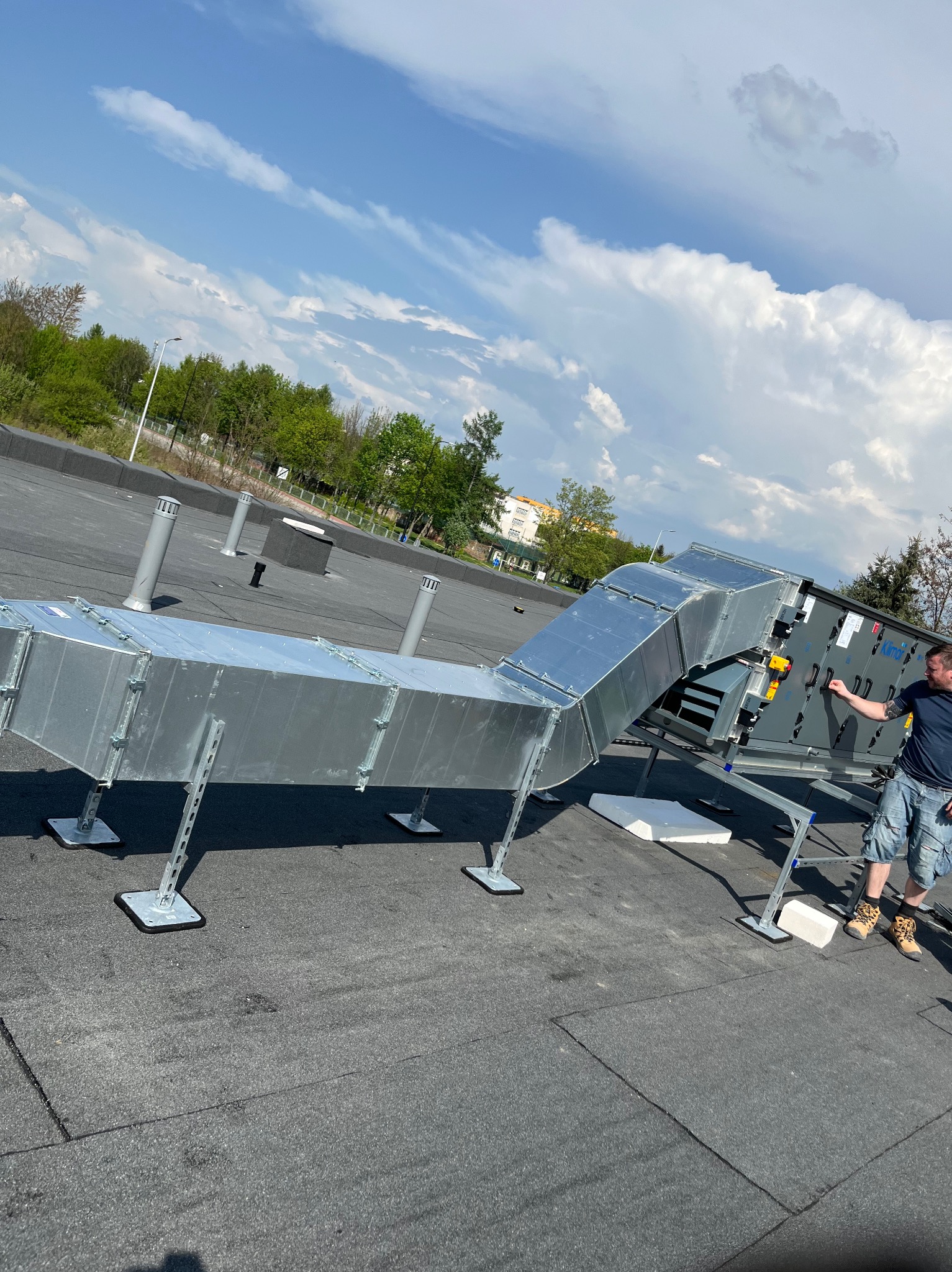 Instalacja wentylacyjna na dachu budynku, widoczny pracownik przy urządzeniu. Metalowe kanały wentylacyjne na wspornikach, błękitne niebo w tle.