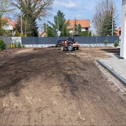 Małgorzata Kalata Usługi Ogrodnicze - Przygotowanie gruntu pod trawnik: mini-traktor wyrównuje ziemię na działce z nasadzeniami przy nowoczesnym domu i szarym ogrodzeniu.