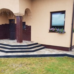 Małgorzata Kalata Usługi Ogrodnicze - Ułożona kostka brukowa przed domem z beżową elewacją i kwiatami w oknach. Schody wejściowe wyłożone płytkami, widoczny trawnik i elementy architektury ogrodowej.