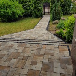 Małgorzata Kalata Usługi Ogrodnicze - Ułożona kostka brukowa w ogrodzie prowadząca do furtki, otoczona zielenią. Widoczne krzewy i trawnik. Kostka w odcieniach brązu i beżu, z czarnymi elementami oddzielającymi.