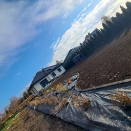 Małgorzata Kalata Usługi Ogrodnicze - Przygotowany teren pod ogród z rozłożoną agrowłókniną, widok na dom i niebo z chmurami. Ukośne ujęcie podkreśla przestrzeń i etap prac.