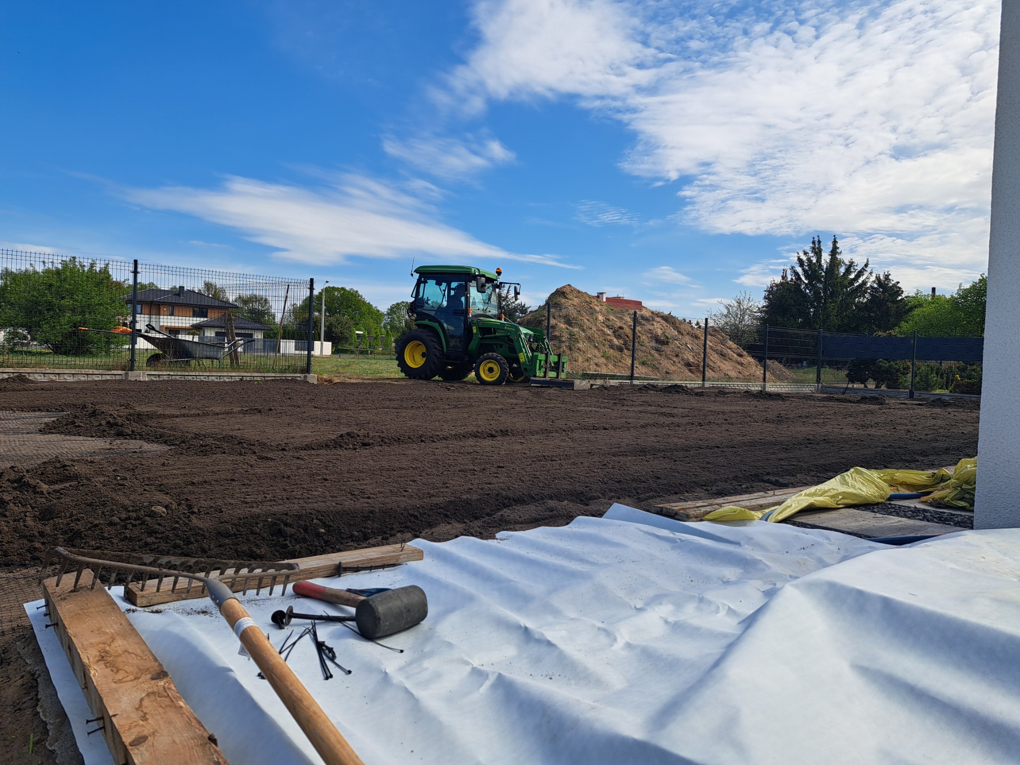 Przygotowany teren pod trawnik z rolki, widoczny traktor, narzędzia ogrodnicze i materiały izolacyjne. W tle ogrodzenie i zabudowa jednorodzinna.