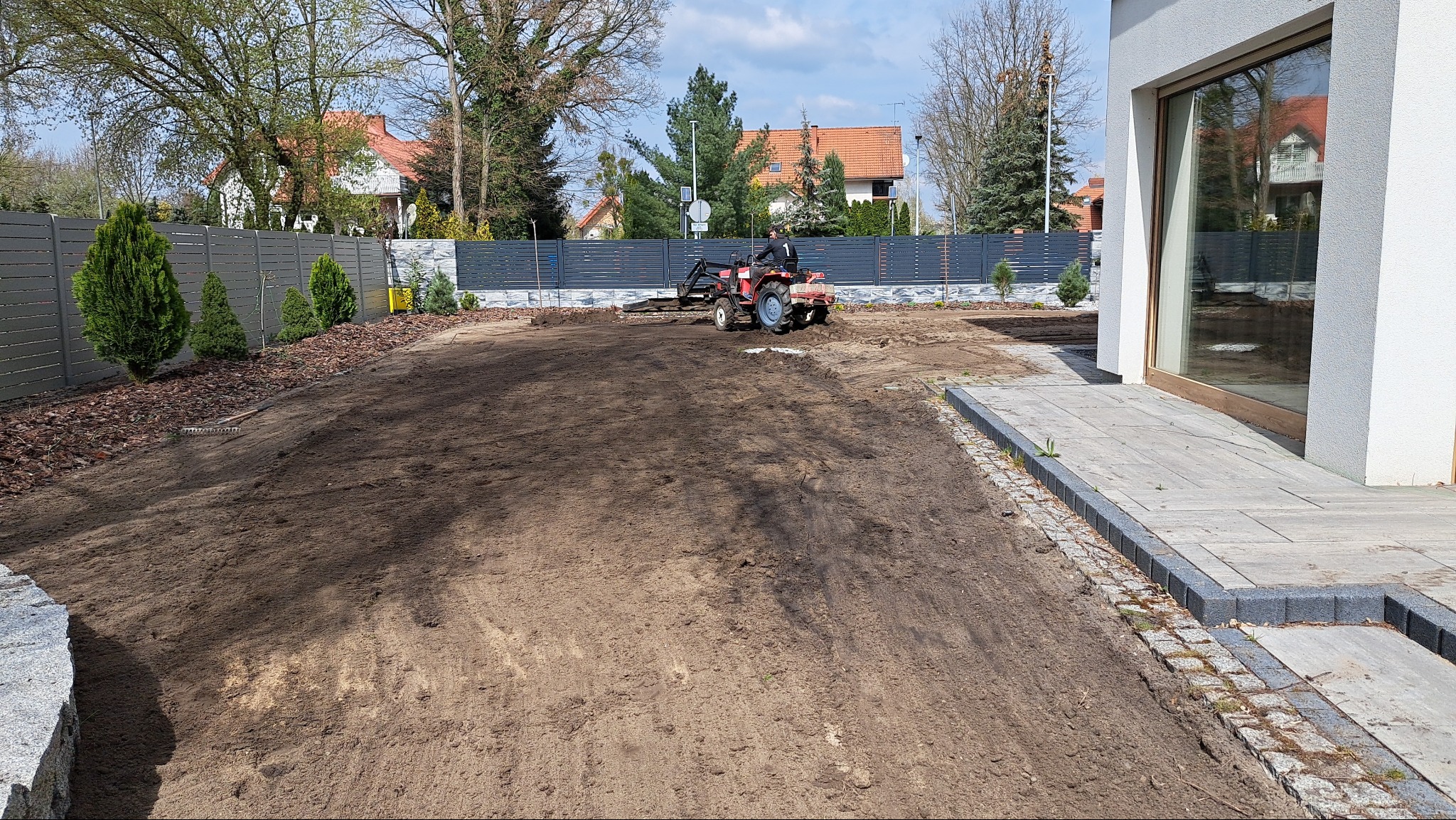 Przygotowanie gruntu pod trawnik: mini-traktor wyrównuje ziemię na działce z nasadzeniami przy nowoczesnym domu i szarym ogrodzeniu.