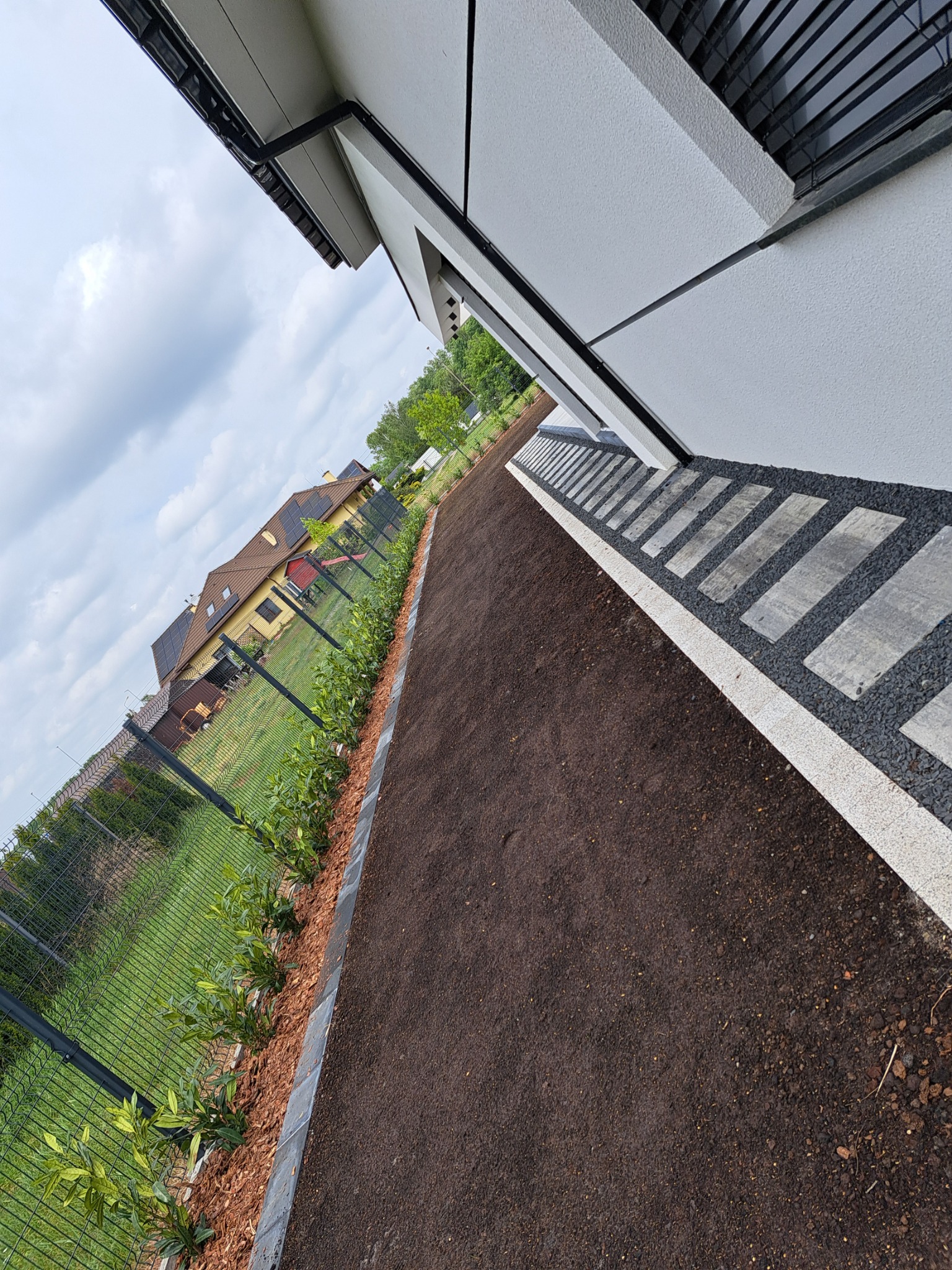 Ścieżka z kostki brukowej i świeżo posadzone rośliny wzdłuż ogrodzenia, widok z boku domu, zieleń trawnika i drzewa w tle, pochmurne niebo.