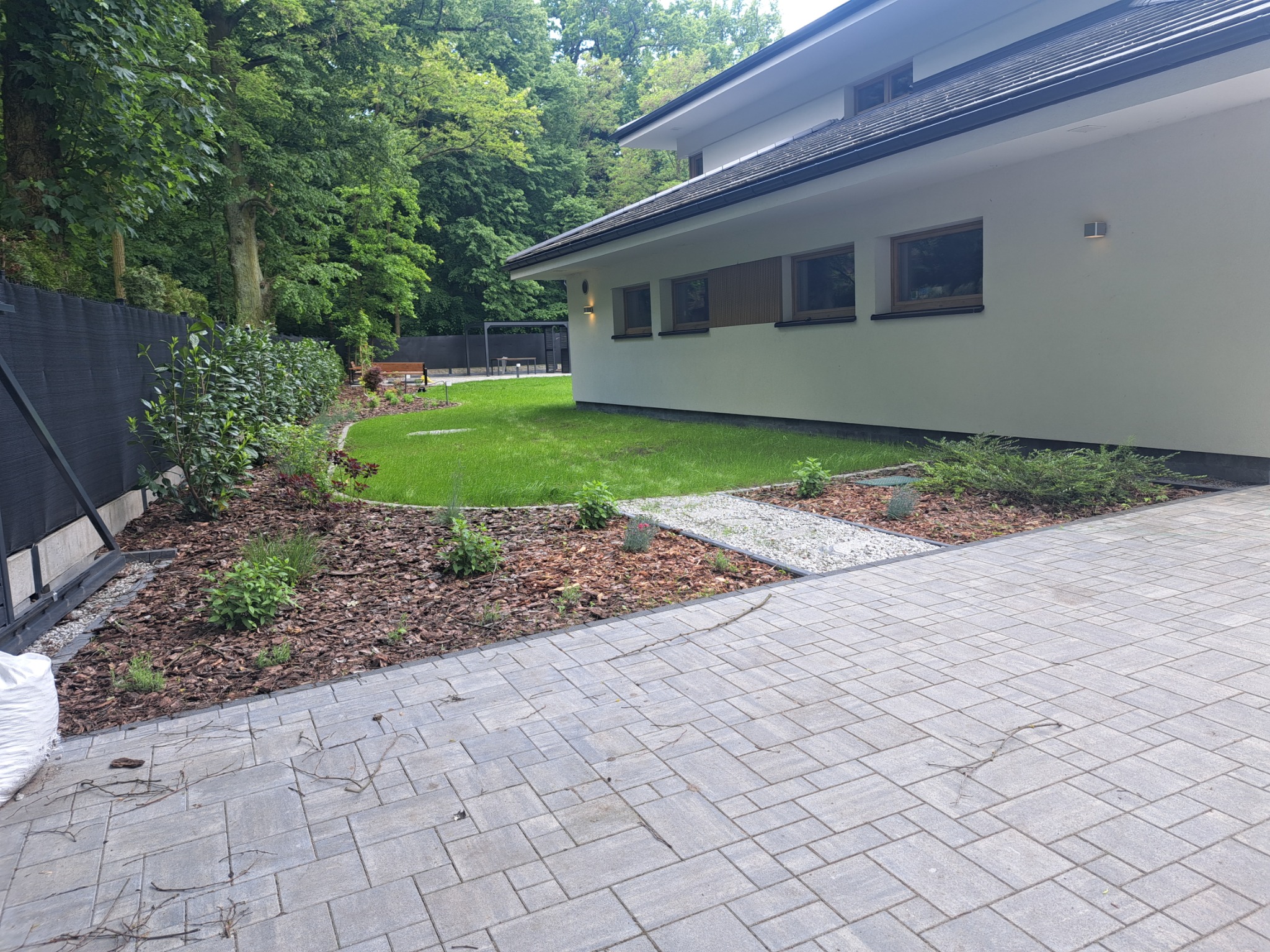 Nowoczesny ogród przydomowy z trawnikiem, rabatami z korą i kamienną ścieżką, widok z brukowanego podjazdu w kierunku domu i zieleni.