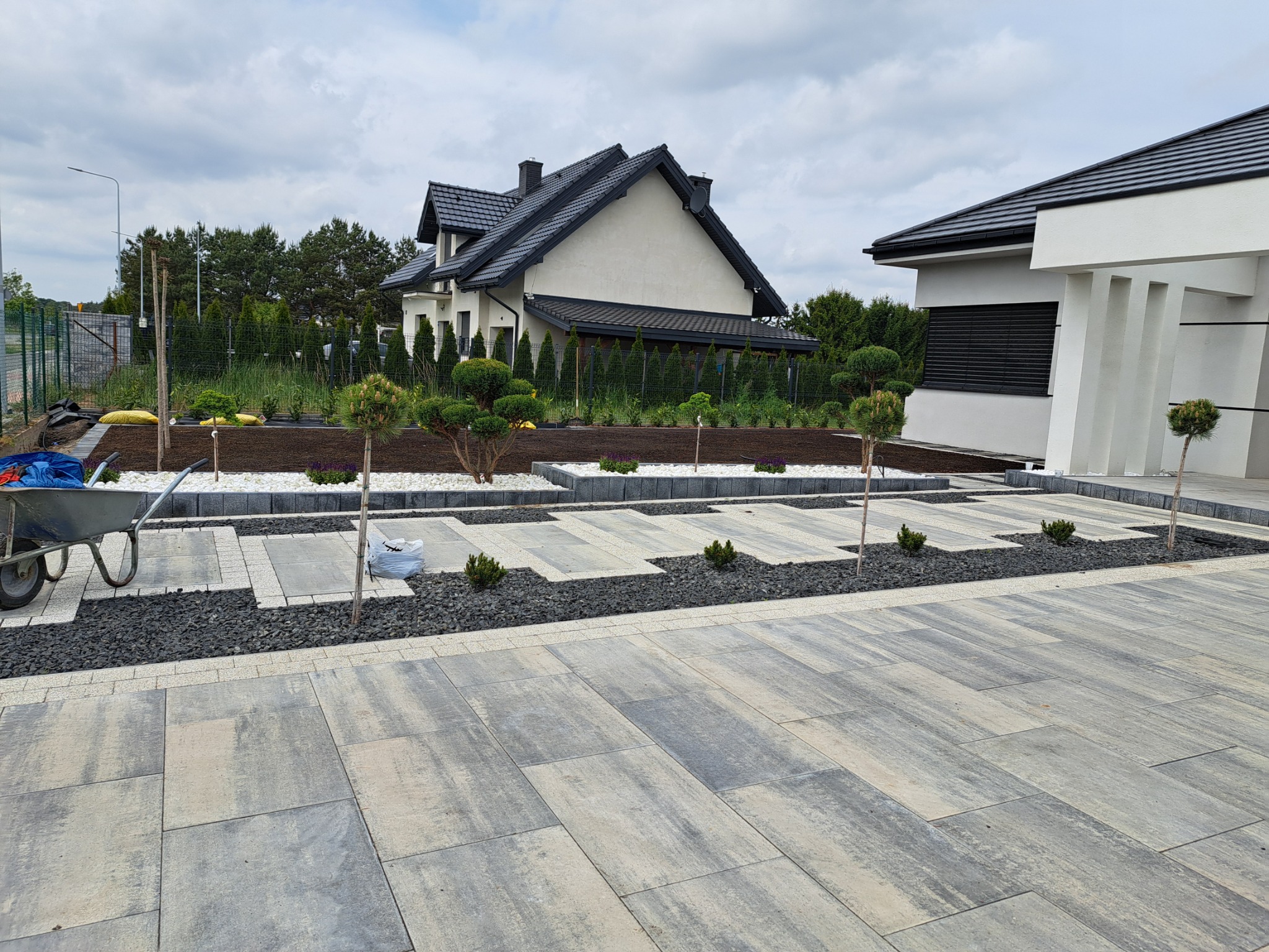 Nowoczesny ogród z szarą kostką brukową, minimalistycznymi rabatami z białym i czarnym grysem, oraz młodymi drzewkami. W tle dom z ciemnym dachem i białą elewacją.