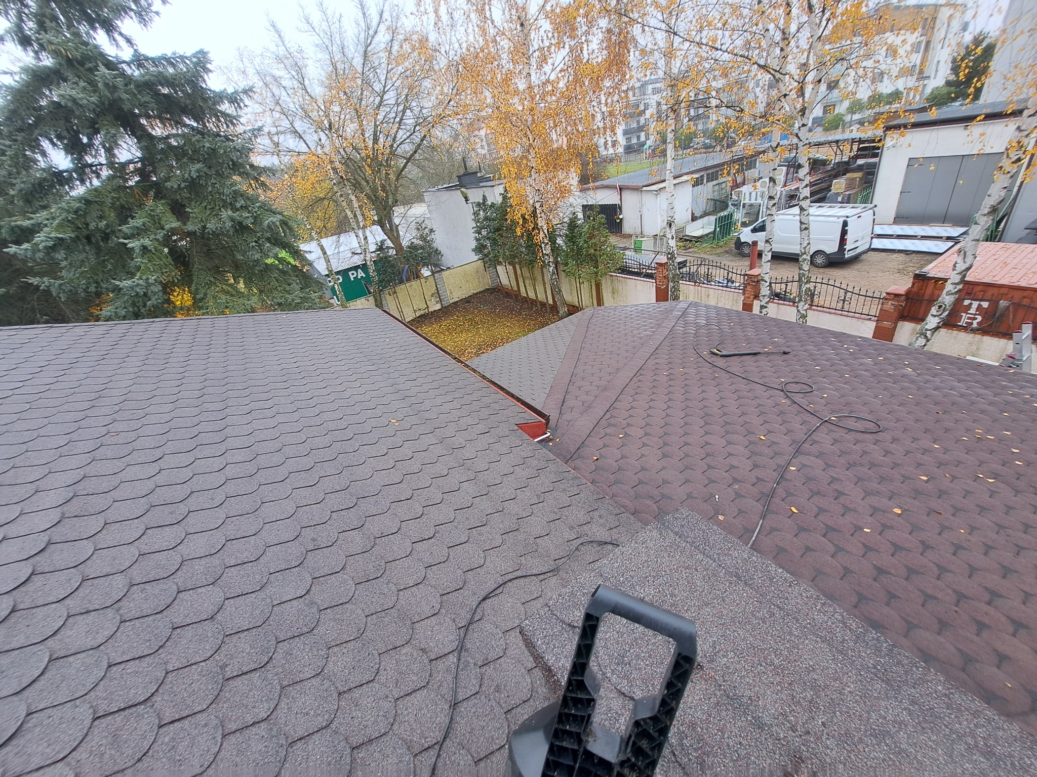 Dach pokryty gontem bitumicznym w odcieniach brązu i szarości, widok z góry na tle drzew i zabudowań. Fragment narzędzia dekarskiego w dolnej części kadru.