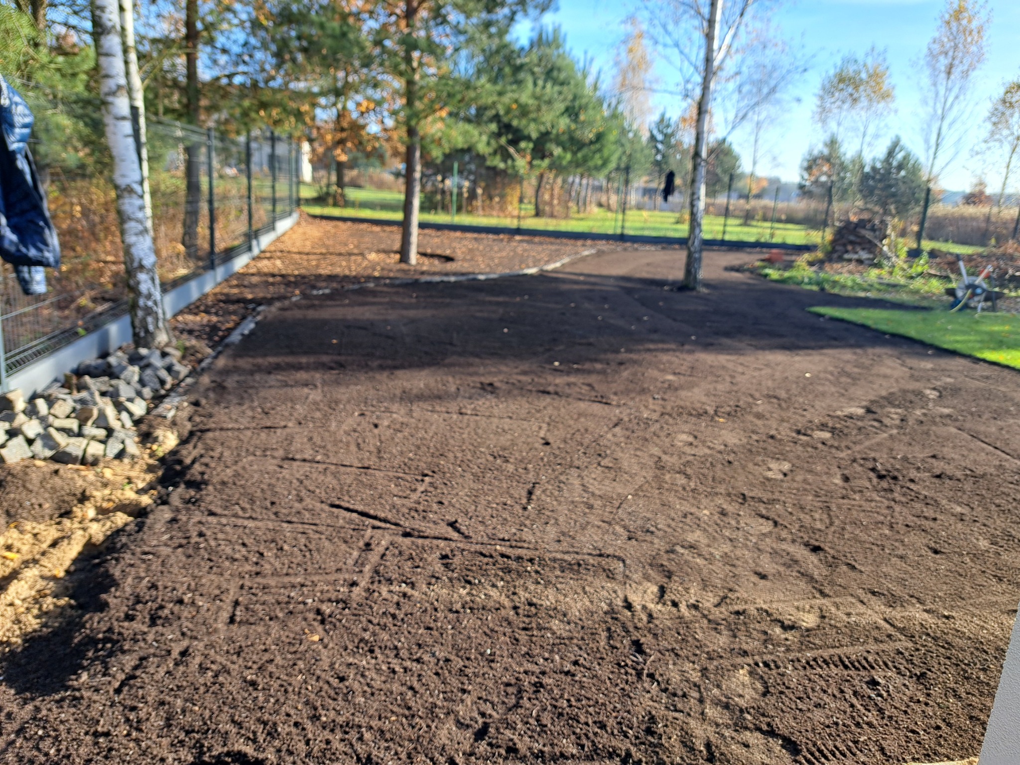 Przygotowany teren pod trawnik z widocznymi śladami po maszynach, obrzeża z kamieni i trawy, drzewa na działce, ogrodzenie panelowe.