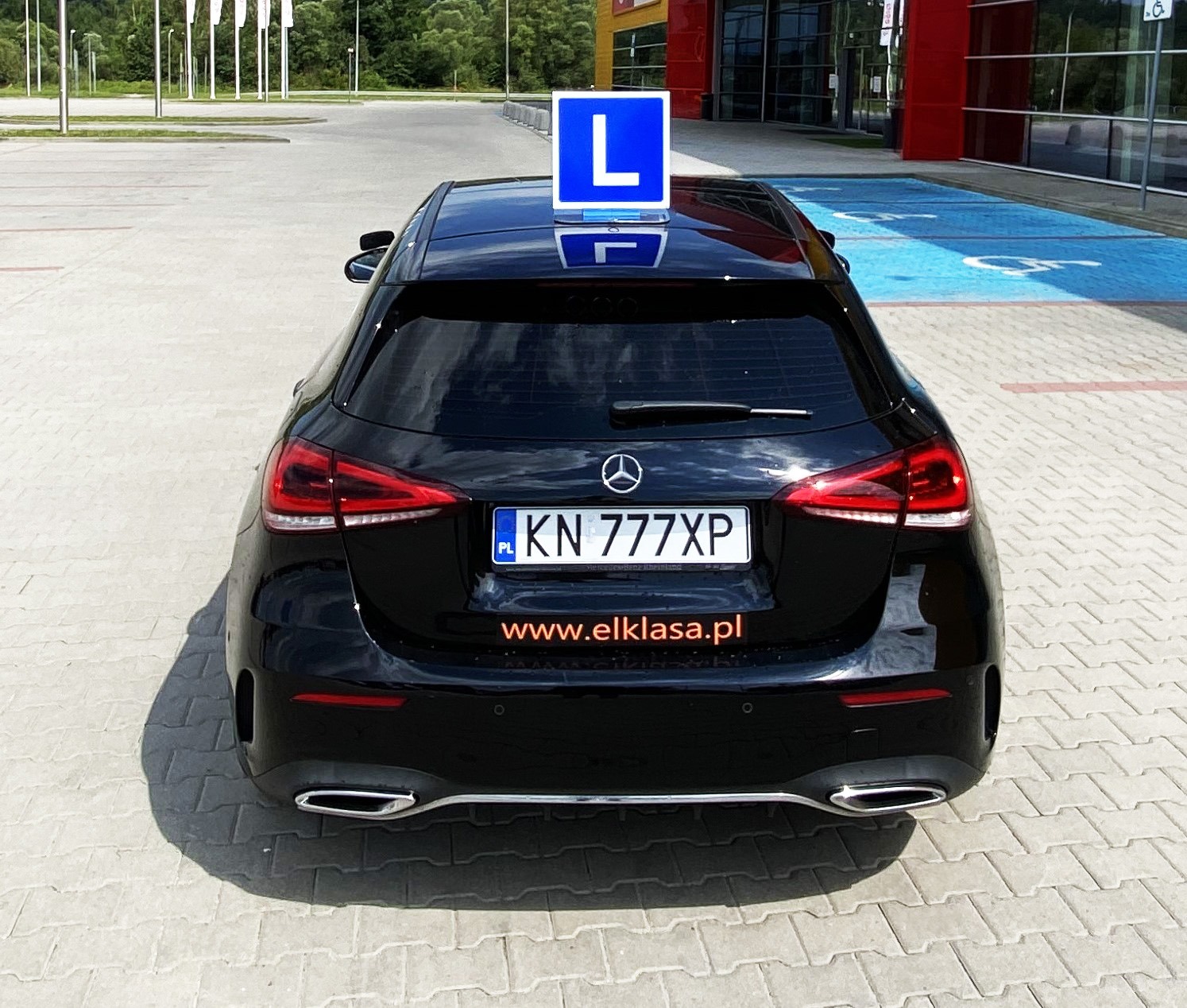 Czarny Mercedes z niebieską tablicą 'L' na dachu, zaparkowany na placu przed budynkiem z czerwonymi elementami.