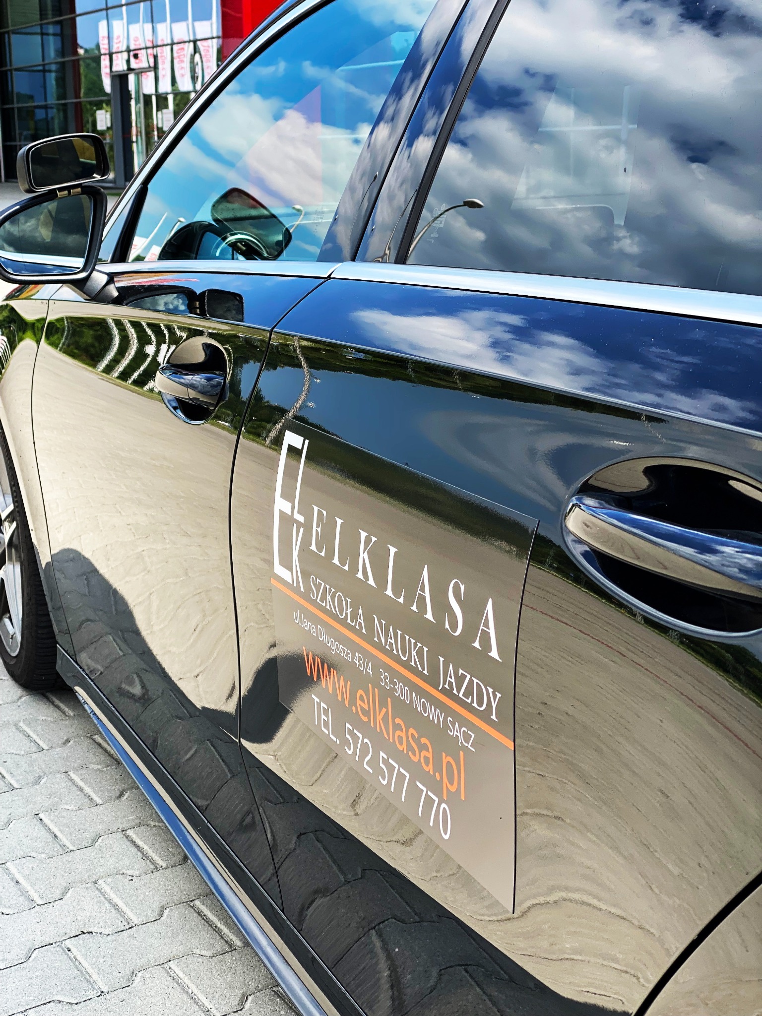 Błyszczący czarny samochód z naklejką informacyjną o szkole nauki jazdy 'Elklasa', zaparkowany na chodniku z kostki brukowej, z odbiciem chmur w karoserii.