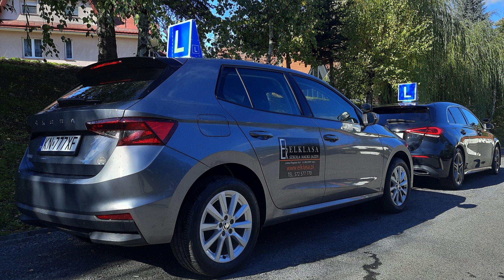 Dwa samochody z niebieskimi znakami 'L' na dachu, zaparkowane obok siebie, szary Skoda Fabia z logo 'Elklasa' z boku.