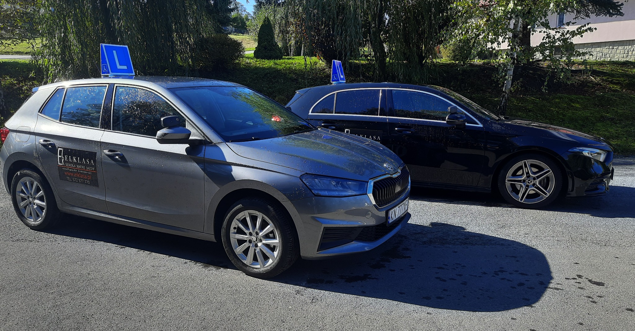 Dwa samochody z literą 'L' na dachu, szary Skoda i czarny Mercedes, zaparkowane na zewnątrz, z widocznym logo i danymi kontaktowymi firmy na boku Skody.