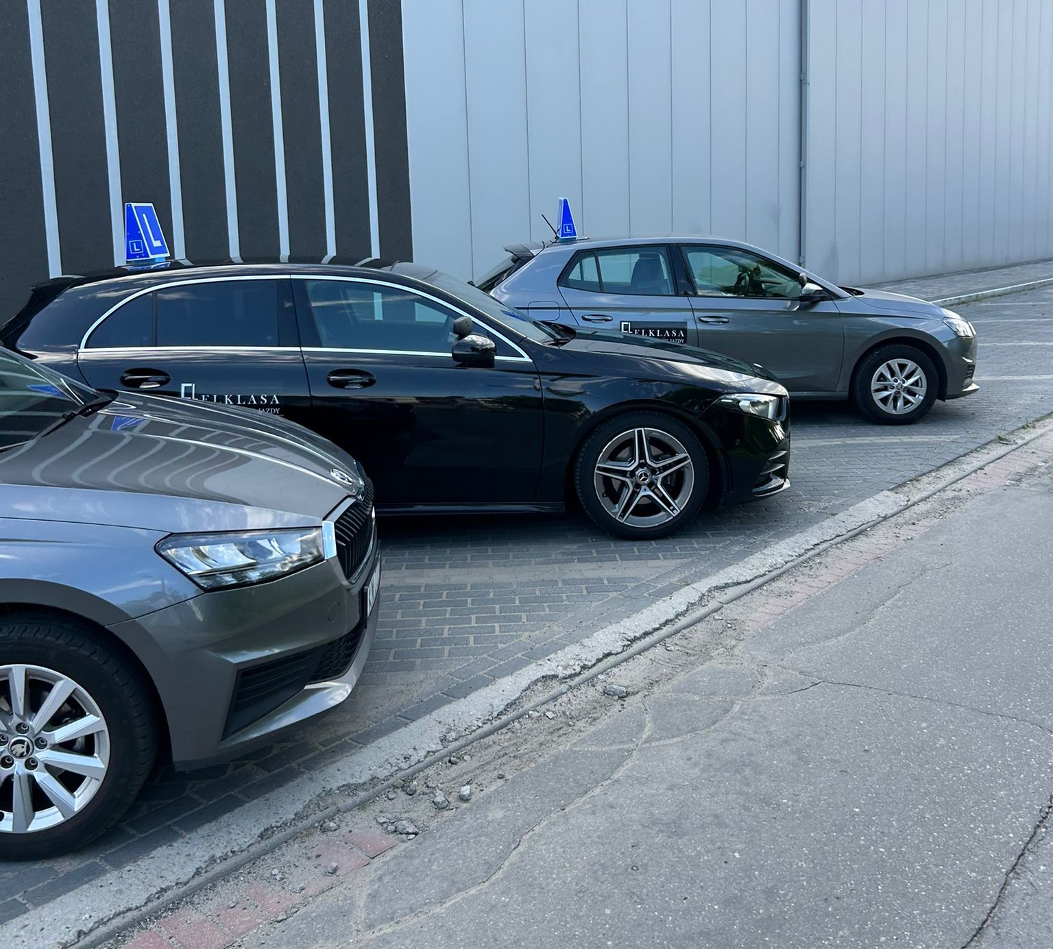 Flota aut oznaczonych symbolem 'L' na dachu, zaparkowana przed nowoczesnym budynkiem. Widoczne modele różnych marek w stonowanych kolorach.