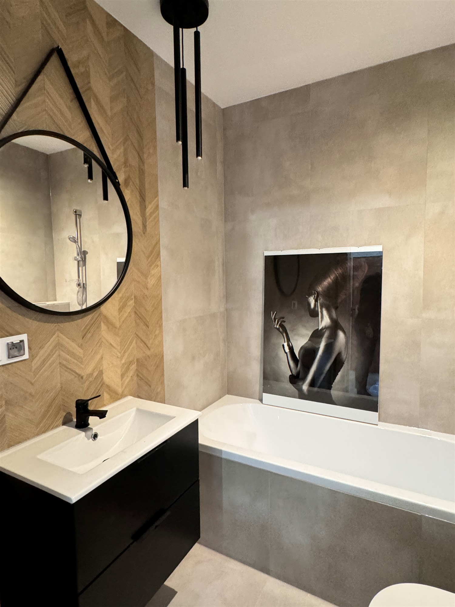 Nowoczesna łazienka z umywalką na czarnej szafce, okrągłym lustrem z czarną ramą, wiszącą lampą z trzema czarnymi tubami i obrazem kobiety nad białą wanną.