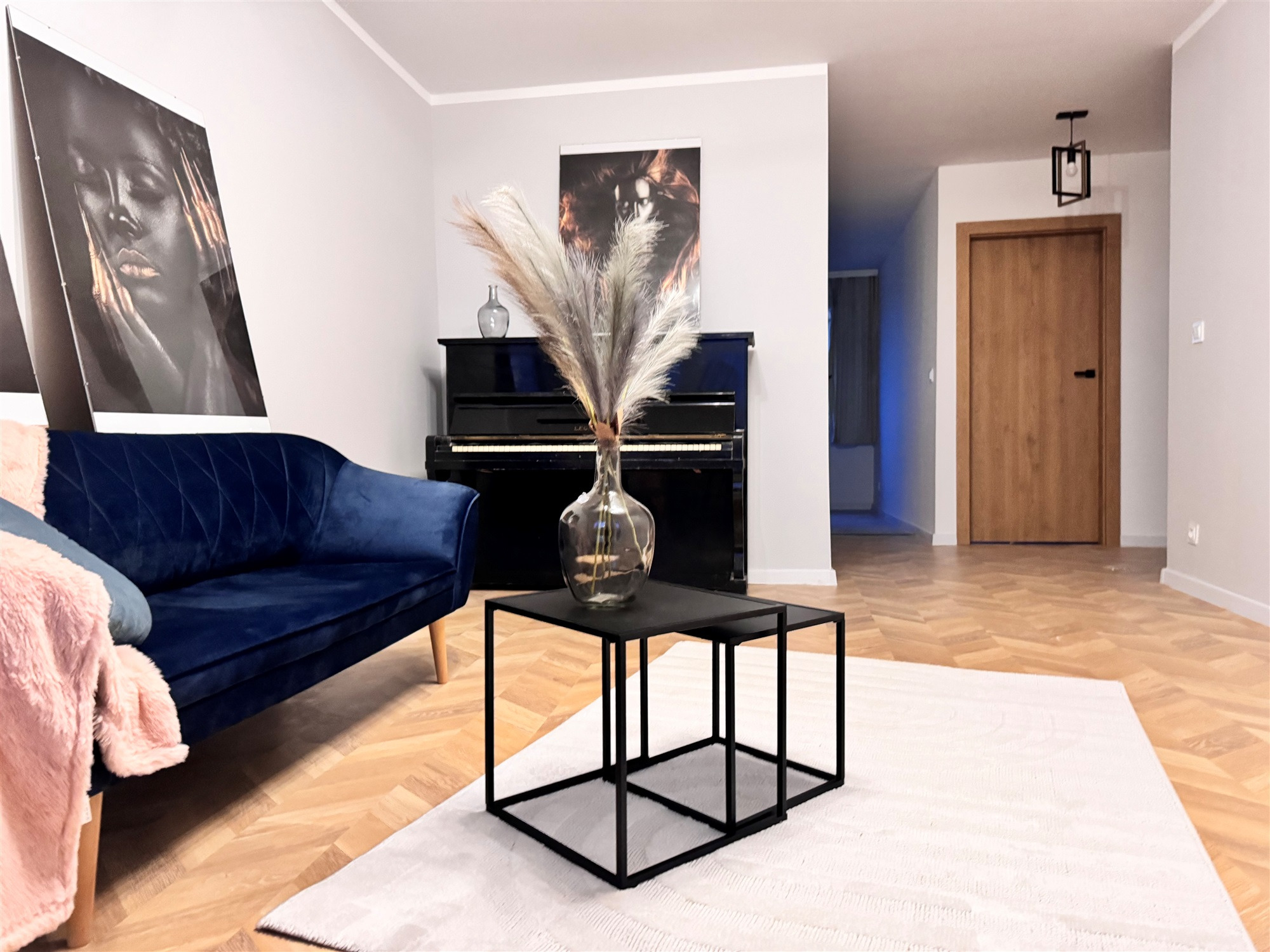 Nowoczesny salon z granatową sofą, czarnym pianinem i abstrakcyjnymi obrazami na ścianie, uzupełniony stolikiem kawowym z metalową ramą i pampasową trawą w szklanym wazonie.