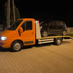 Pomarańczowa ciężarówka z platformą transportową przewozi czarny samochód osobowy na tle nocnego krajobrazu z ogrodzeniem i drzewami.