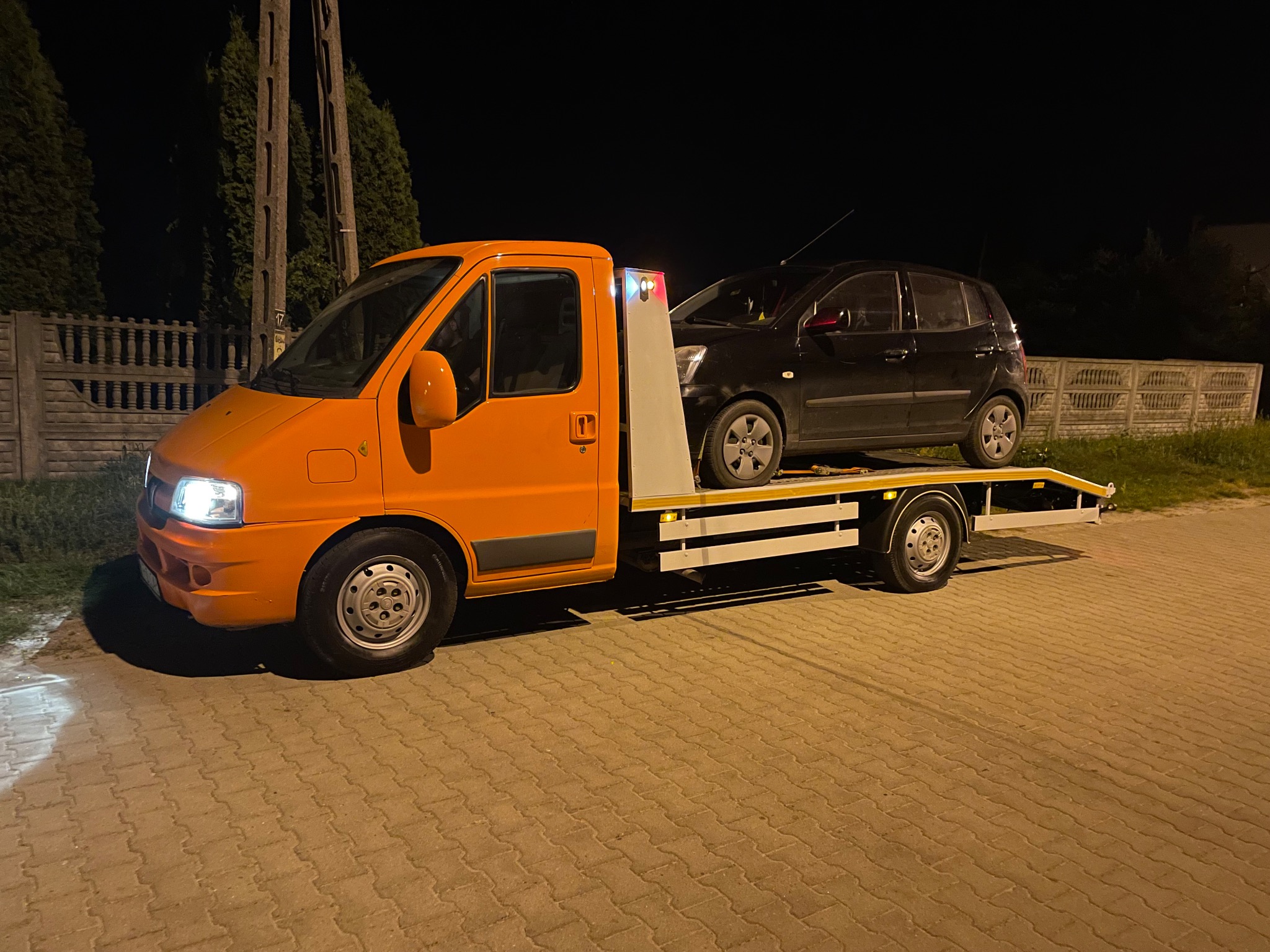 Pomarańczowa ciężarówka z platformą transportową przewozi czarny samochód osobowy na tle nocnego krajobrazu z ogrodzeniem i drzewami.
