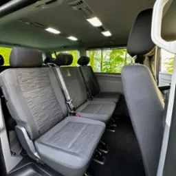 Wnętrze przestronnego busa pasażerskiego z trzema rzędami szarych foteli z teksturowaną tapicerką, widok z boku na czarną podłogę i zielone drzewa za oknem.