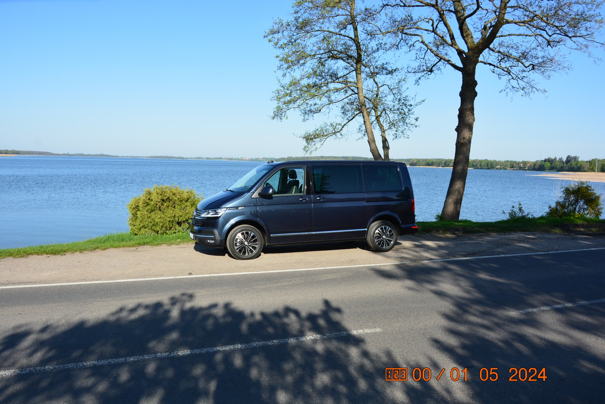 Granatowy bus Volkswagen Transporter zaparkowany na poboczu drogi z widokiem na jezioro i drzewa w słoneczny dzień.