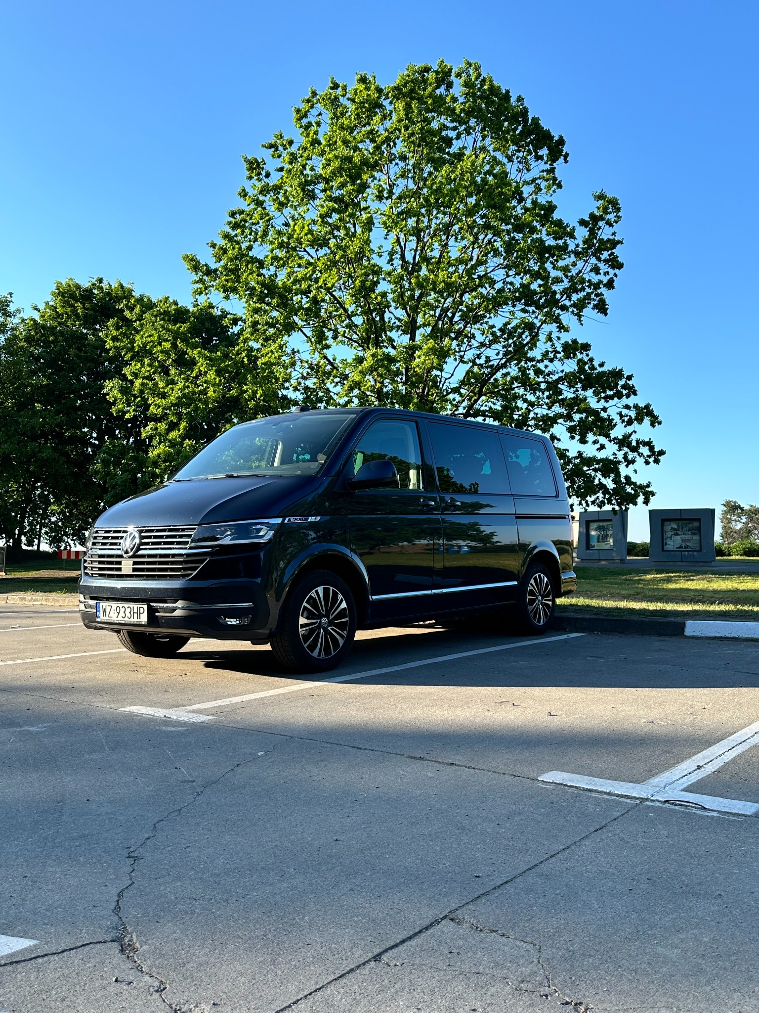 Czarny Volkswagen Transporter T6.1 zaparkowany na parkingu z zielonymi drzewami i pomnikami w tle, widoczne pęknięcia na asfalcie.