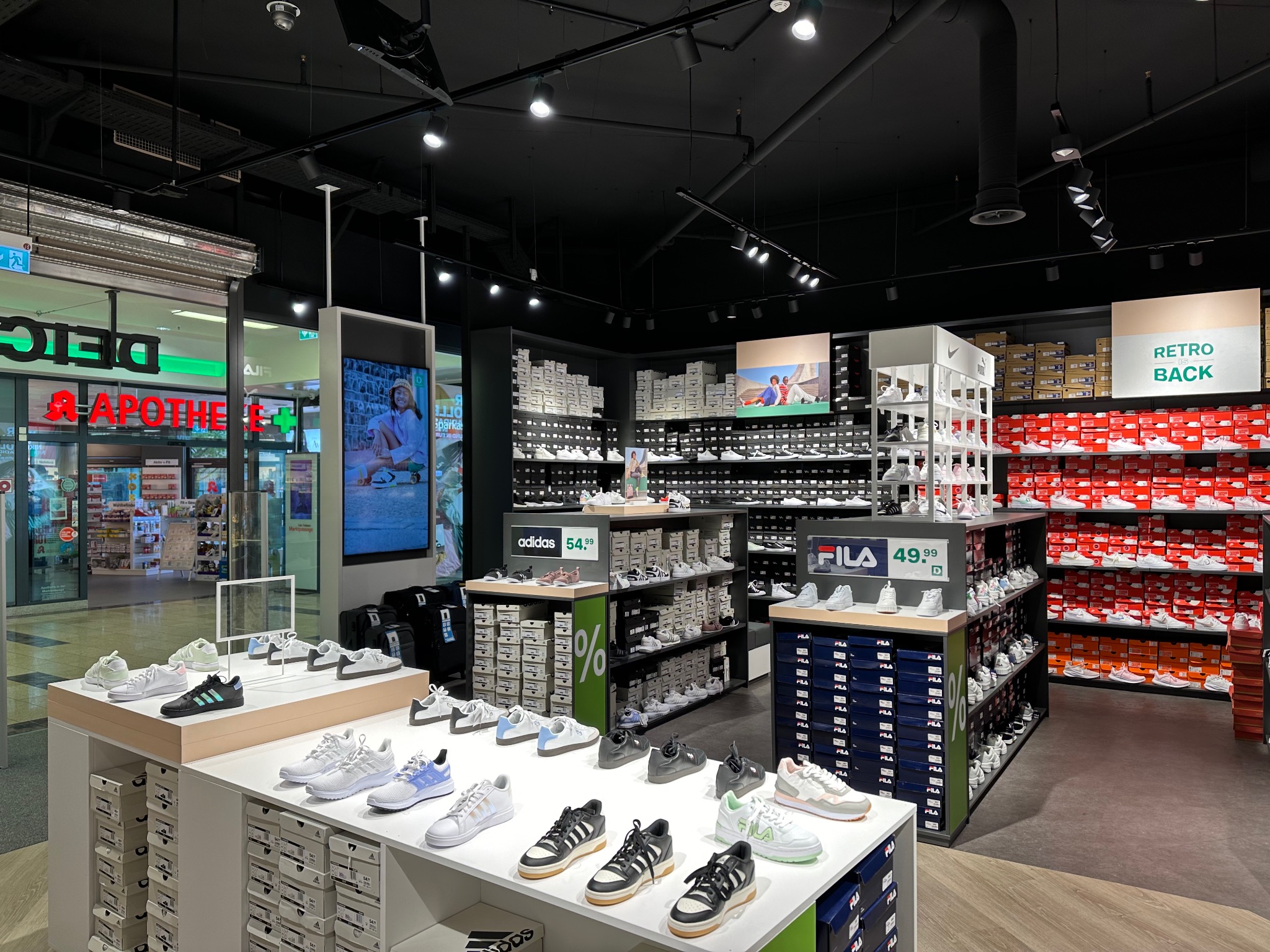 Wnętrze sklepu obuwniczego z ekspozycją butów sportowych marek Adidas i Fila na białych regałach i półkach, z widocznymi cenami i pudełkami. Oświetlenie szynowe sufitowe. W tle widoczna witryna...