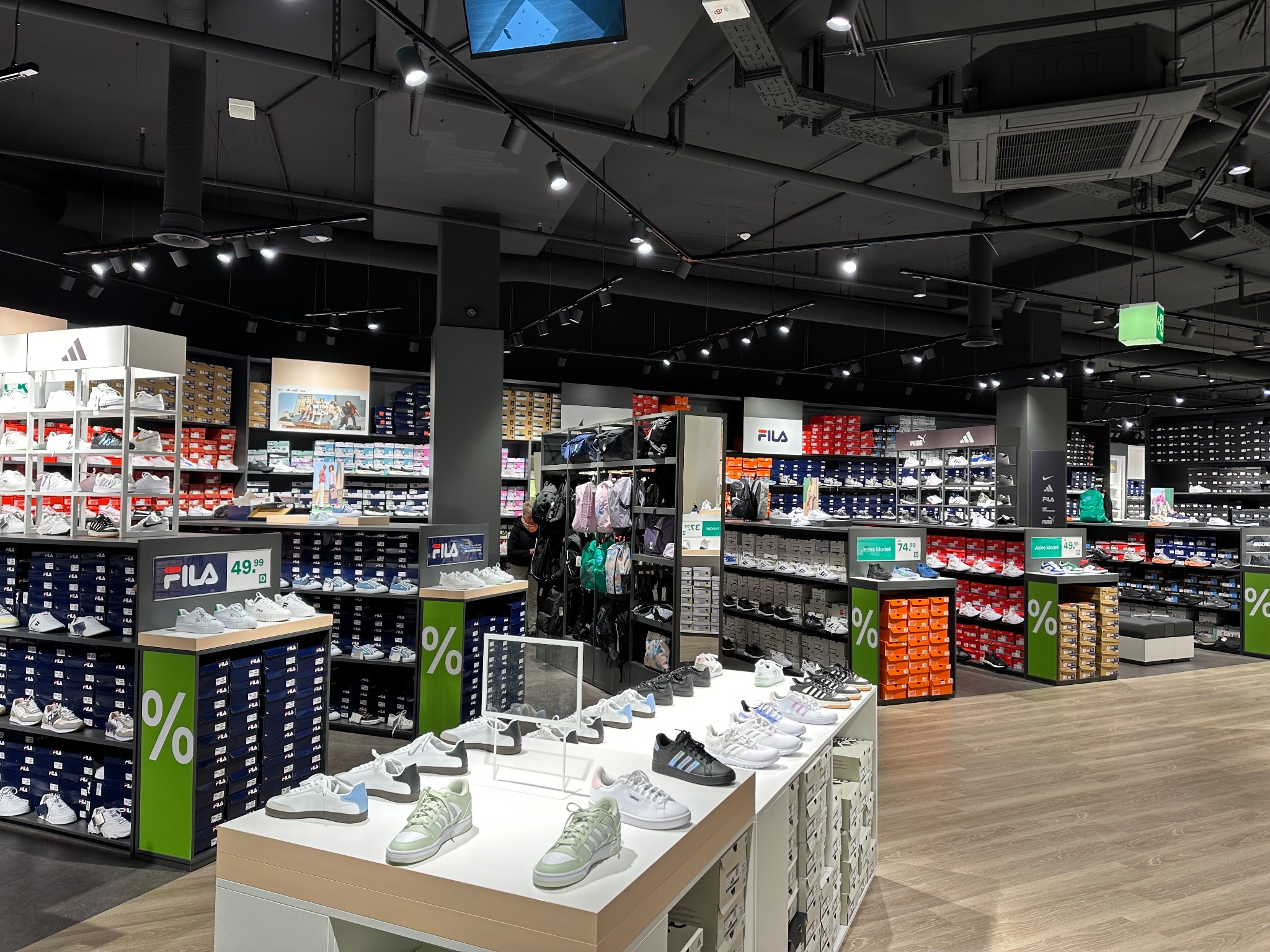 Wnętrze sklepu obuwniczego w Poznaniu, z widocznym oświetleniem punktowym na suficie, ekspozycją obuwia sportowego marki Fila i Adidas na regałach i stołach oraz elementami wentylacji.