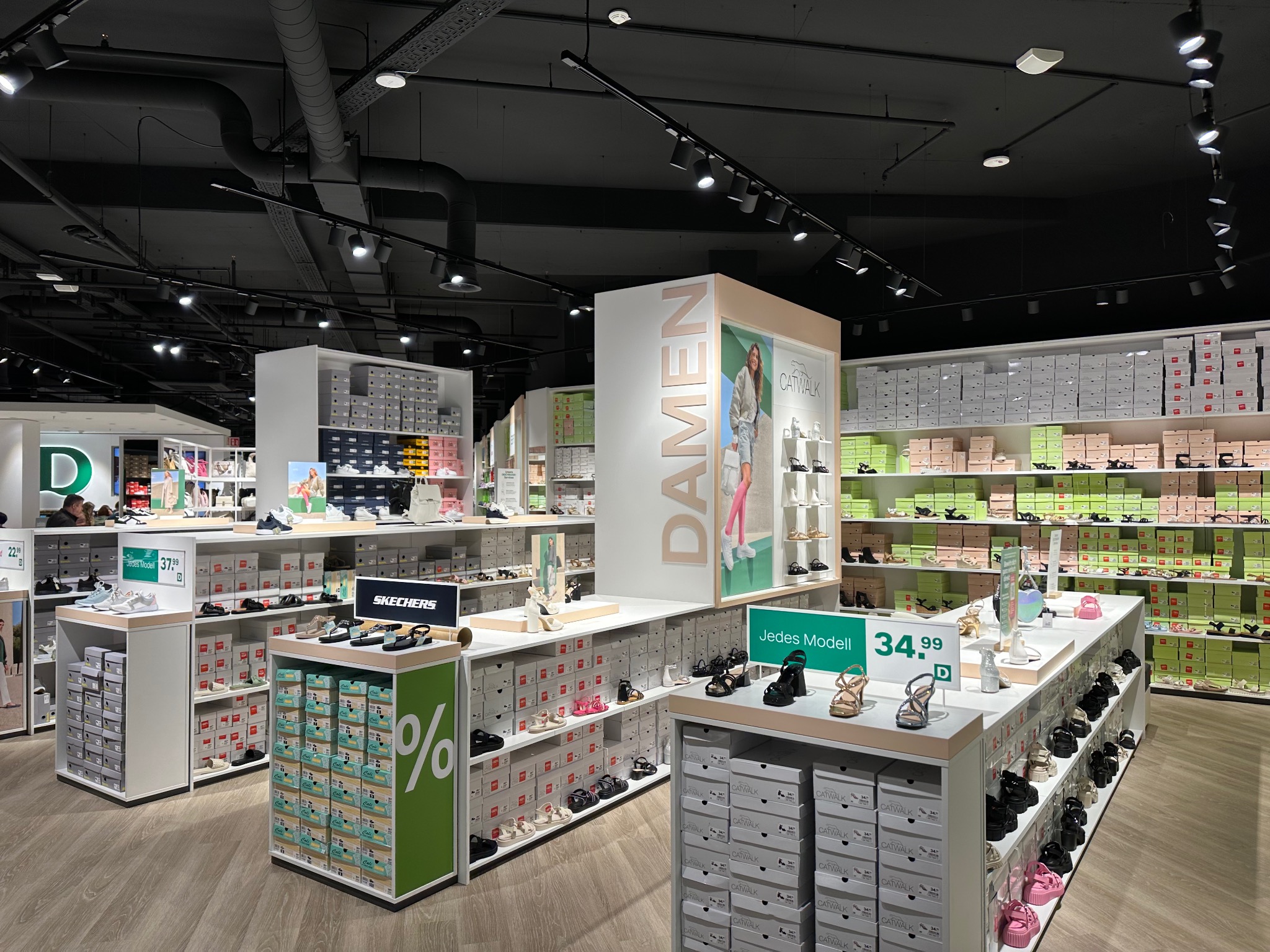 Oświetlenie szynowe w sklepie obuwniczym z ekspozycją obuwia na półkach i wyspach, widoczne pudełka z butami i promocje cenowe.