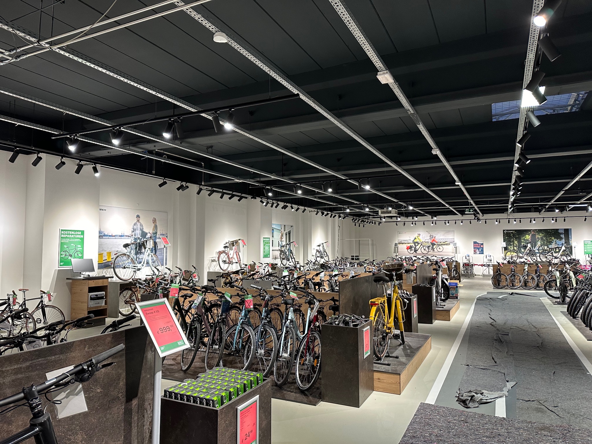 Wnętrze sklepu rowerowego z ekspozycją rowerów różnych typów i cen, oświetlone reflektorami szynowymi na czarnym suficie podwieszanym.