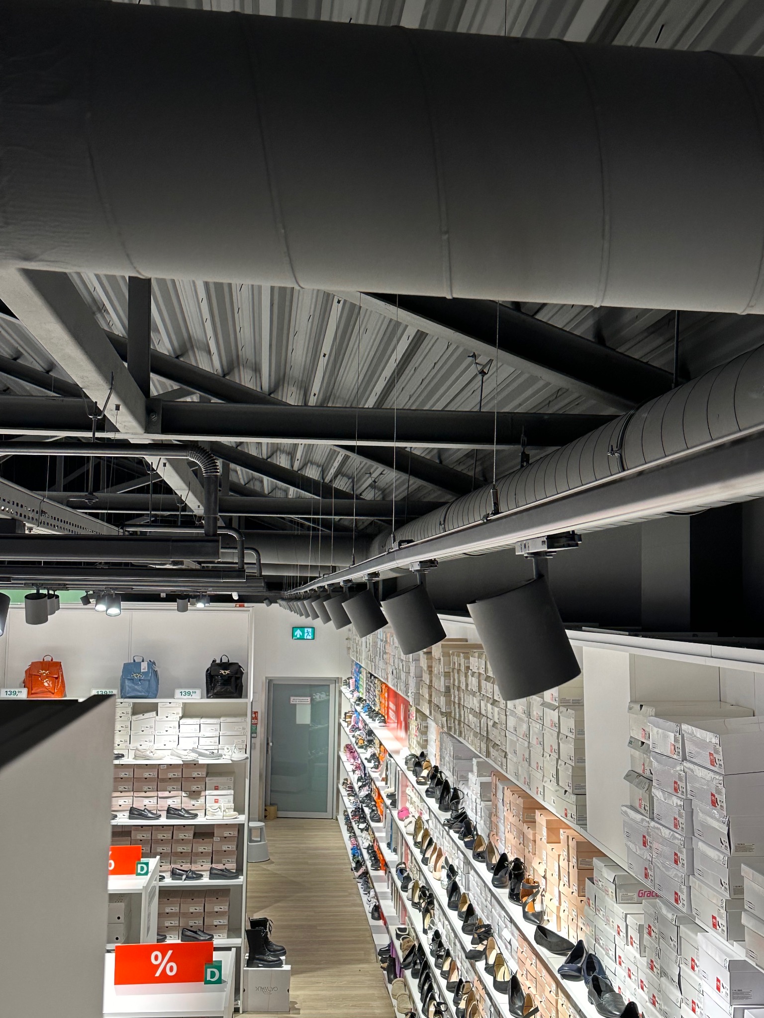 Wnętrze sklepu obuwniczego z widocznymi instalacjami pod sufitem, w tym szynami i reflektorami oświetleniowymi, oraz rzędami półek z butami i pudełkami, ukazując układ sklepu i system oświetlenia.