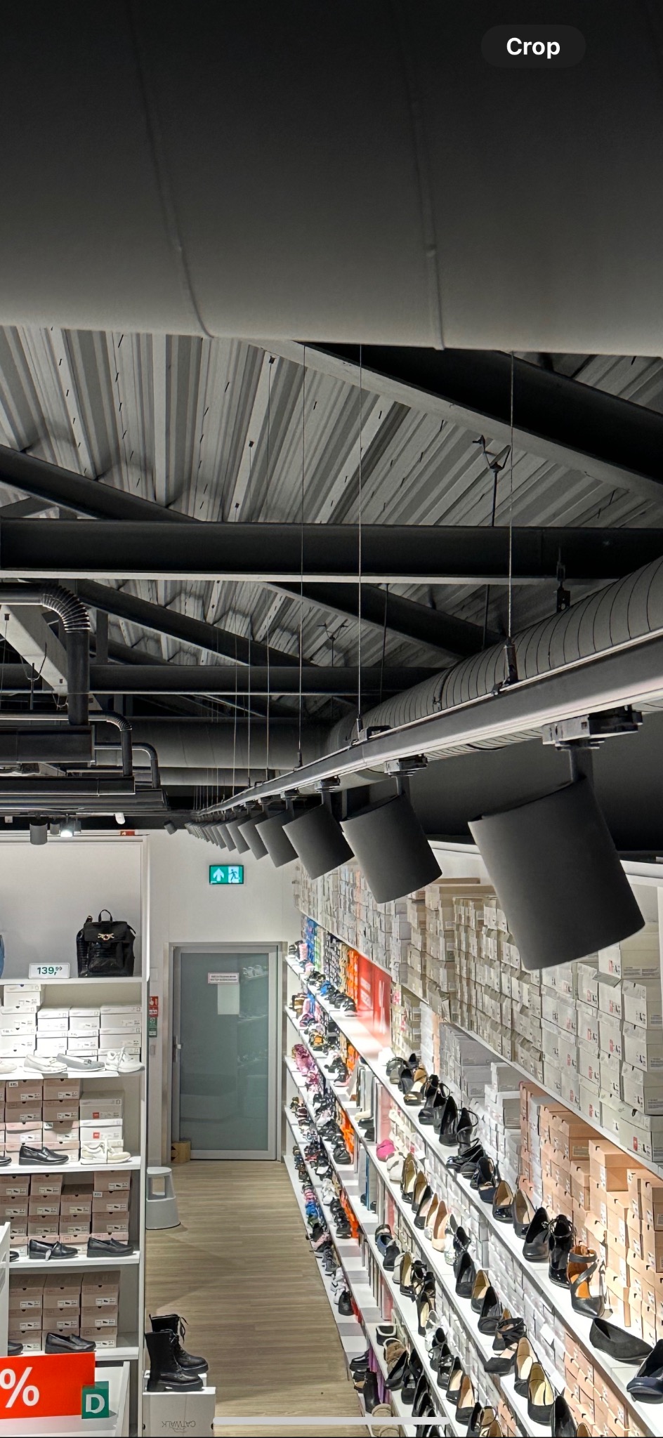 Rząd czarnych reflektorów szynowych oświetla półki z butami w sklepie, widoczne rury wentylacyjne pod sufitem i znak ewakuacyjny.