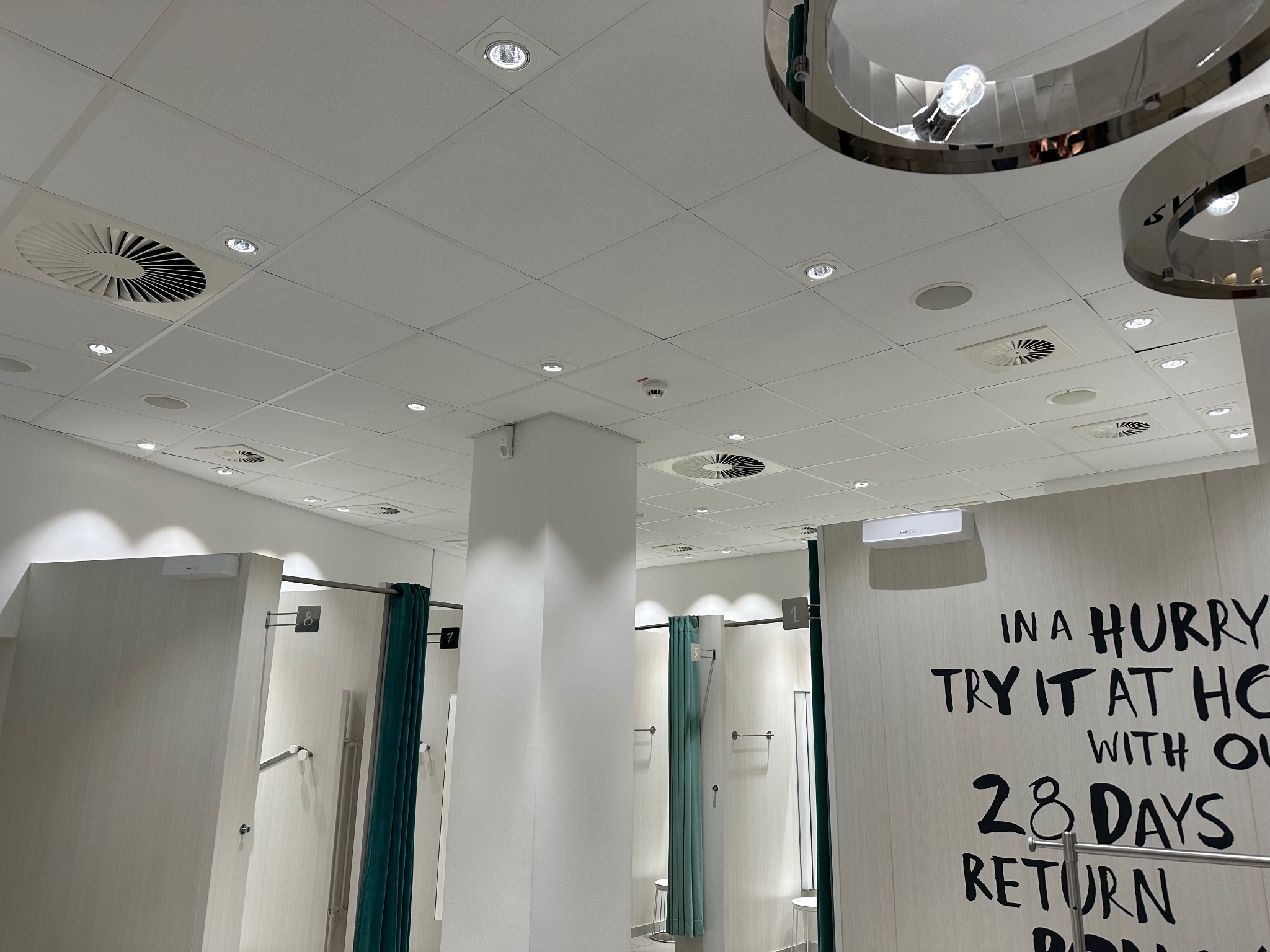 Sufit w przymierzalni sklepu z widocznymi punktowymi oprawami oświetleniowymi, okrągłymi lampami dekoracyjnymi i kratkami wentylacyjnymi.