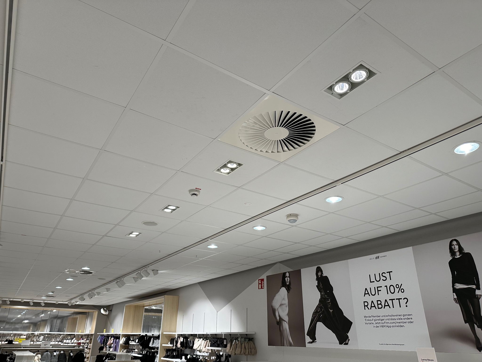 Sufit podwieszany w sklepie z widocznymi elementami instalacji: okrągły dyfuzor wentylacyjny, reflektory sufitowe LED wpuszczane w kwadratowych oprawach oraz okrągłe czujniki dymu.