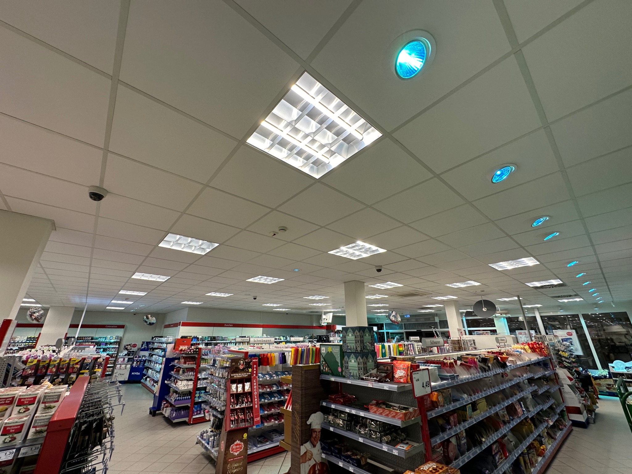 Sufit sklepu z panelami oświetleniowymi LED i punktowymi lampami halogenowymi, widoczne kamery przemysłowe.