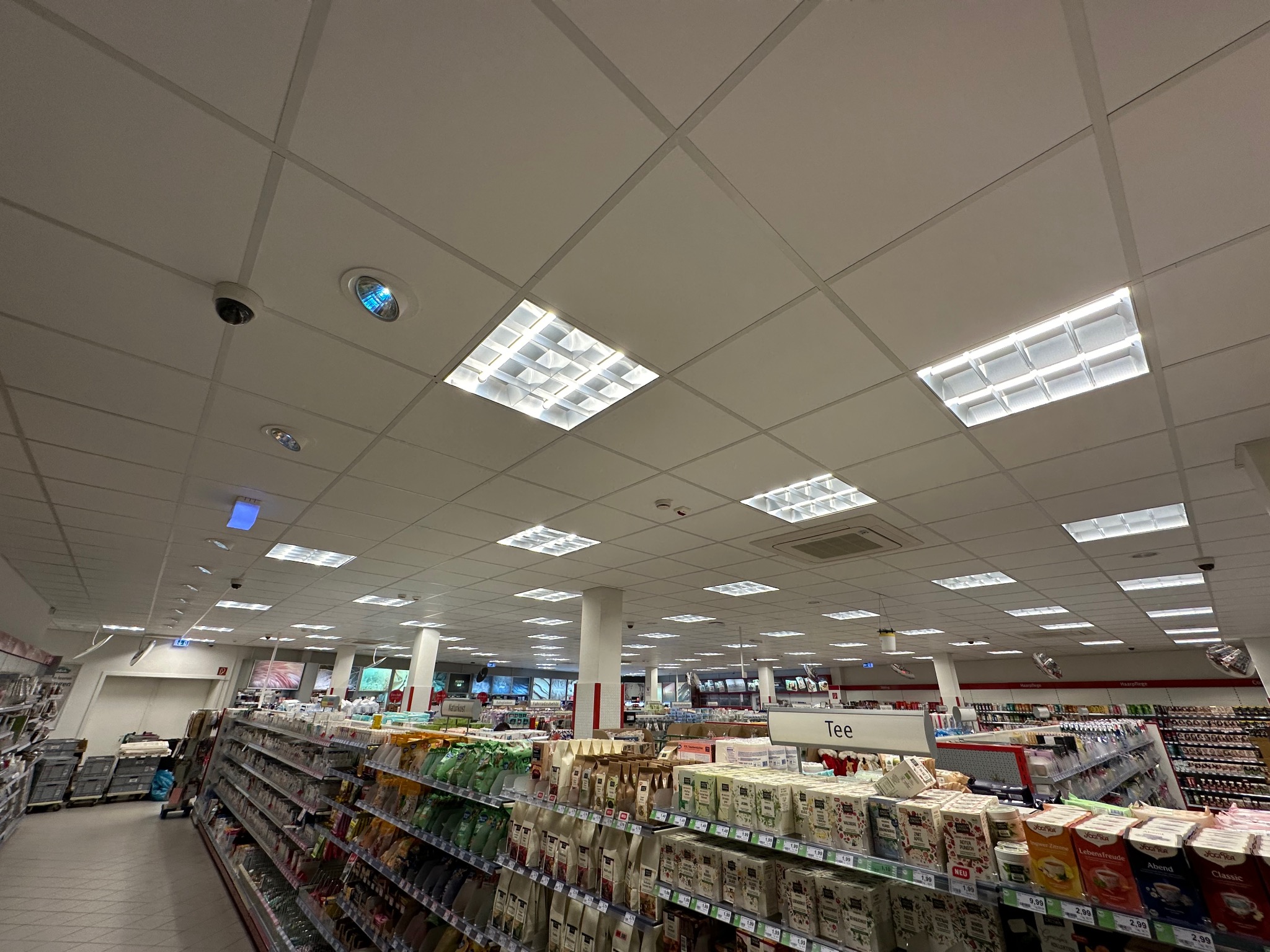 Sufit w supermarkecie z rastrowymi lampami, kamerami monitoringu i systemem wentylacji, widok z dołu na oświetlone wnętrze sklepu z półkami pełnymi towarów.