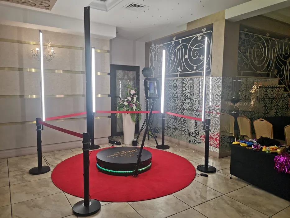 Rozstawiona fotobudka 360 na czerwonym dywanie, otoczona barierkami z czerwonej taśmy i oświetleniem LED, w tle dekoracyjna ściana i stolik z rekwizytami.