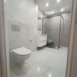 Nowoczesna łazienka z marmurowymi płytkami, wiszącą toaletą, szafką umywalkową i kabiną prysznicową z matowego szkła, widoczna zza uchylonych drzwi.