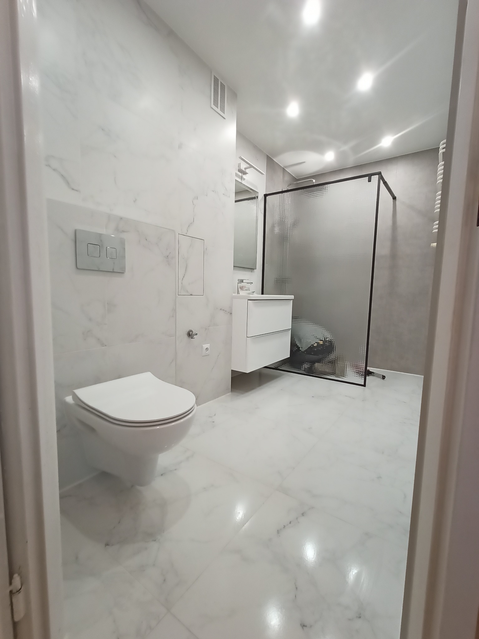 Nowoczesna łazienka z marmurowymi płytkami, wiszącą toaletą, szafką umywalkową i kabiną prysznicową z matowego szkła, widoczna zza uchylonych drzwi.