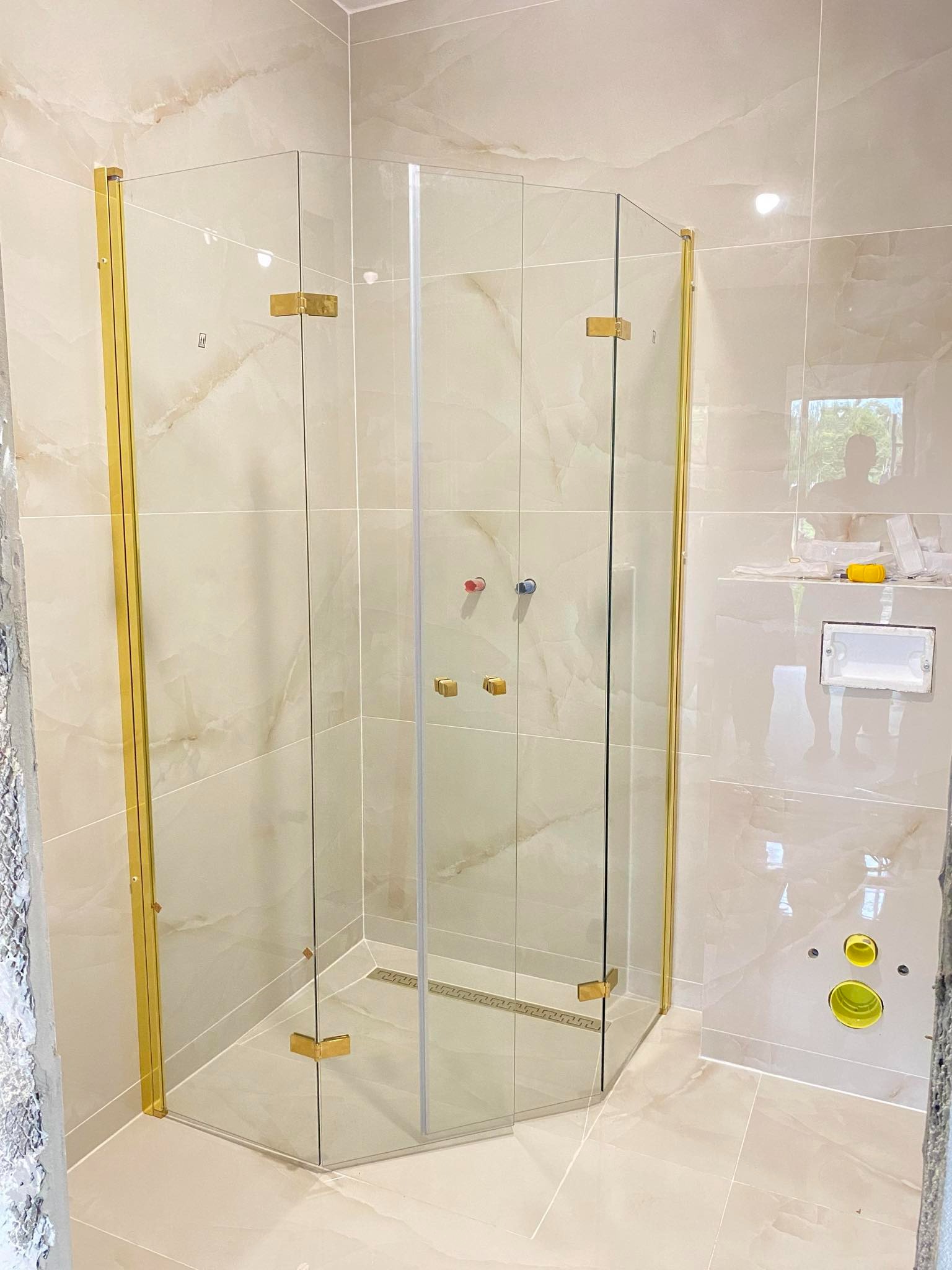 Narożna kabina prysznicowa ze złotymi okuciami, zamontowana w łazience wykończonej beżowymi płytkami imitującymi marmur, widoczny odpływ liniowy i przygotowane przyłącza hydrauliczne.